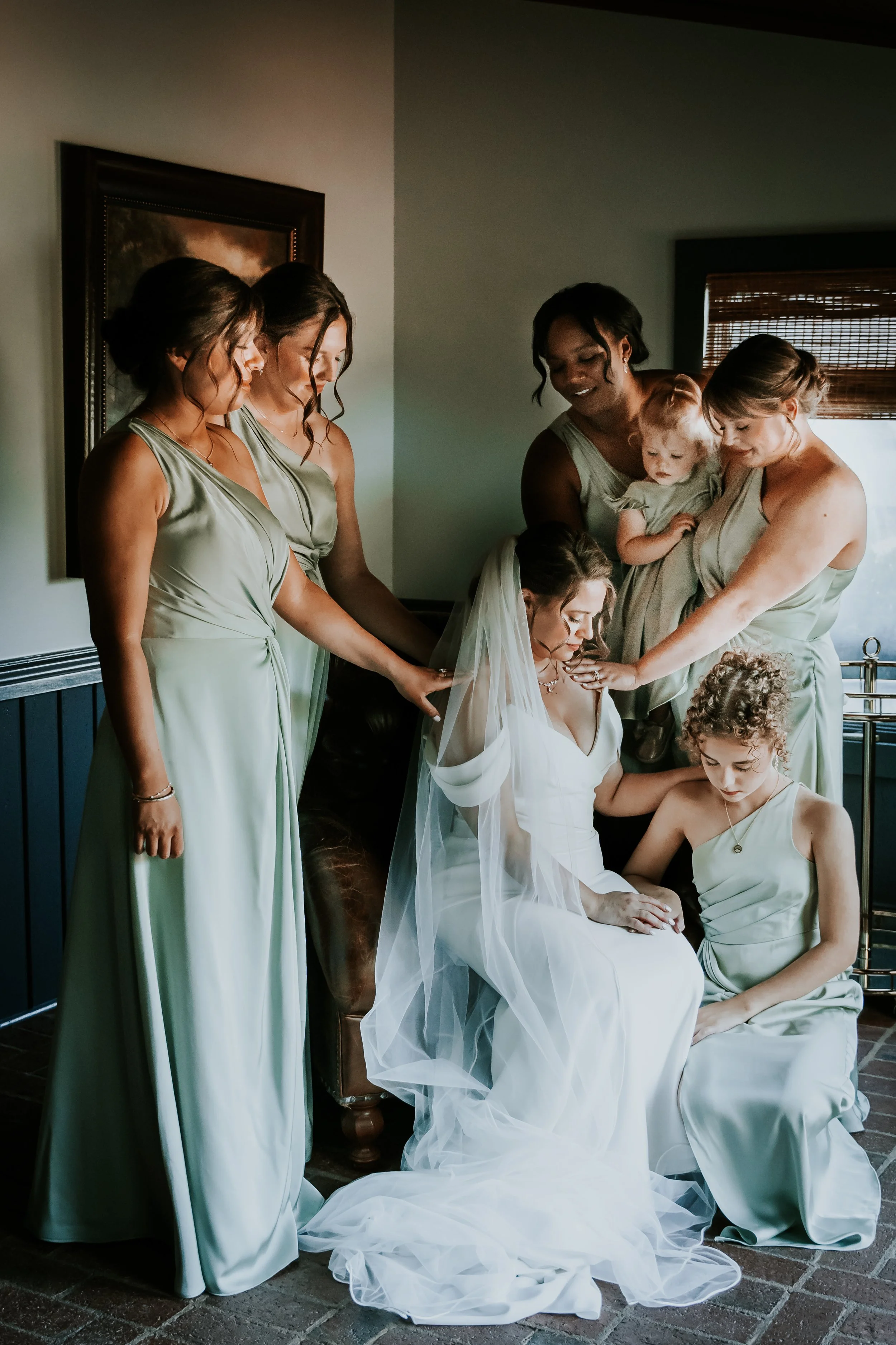 A bride sitting on a chair with her wedding dress for her wedding surrounded by five bridesmaids in green dresses, with one holding a small child, all gathered around her in a room.