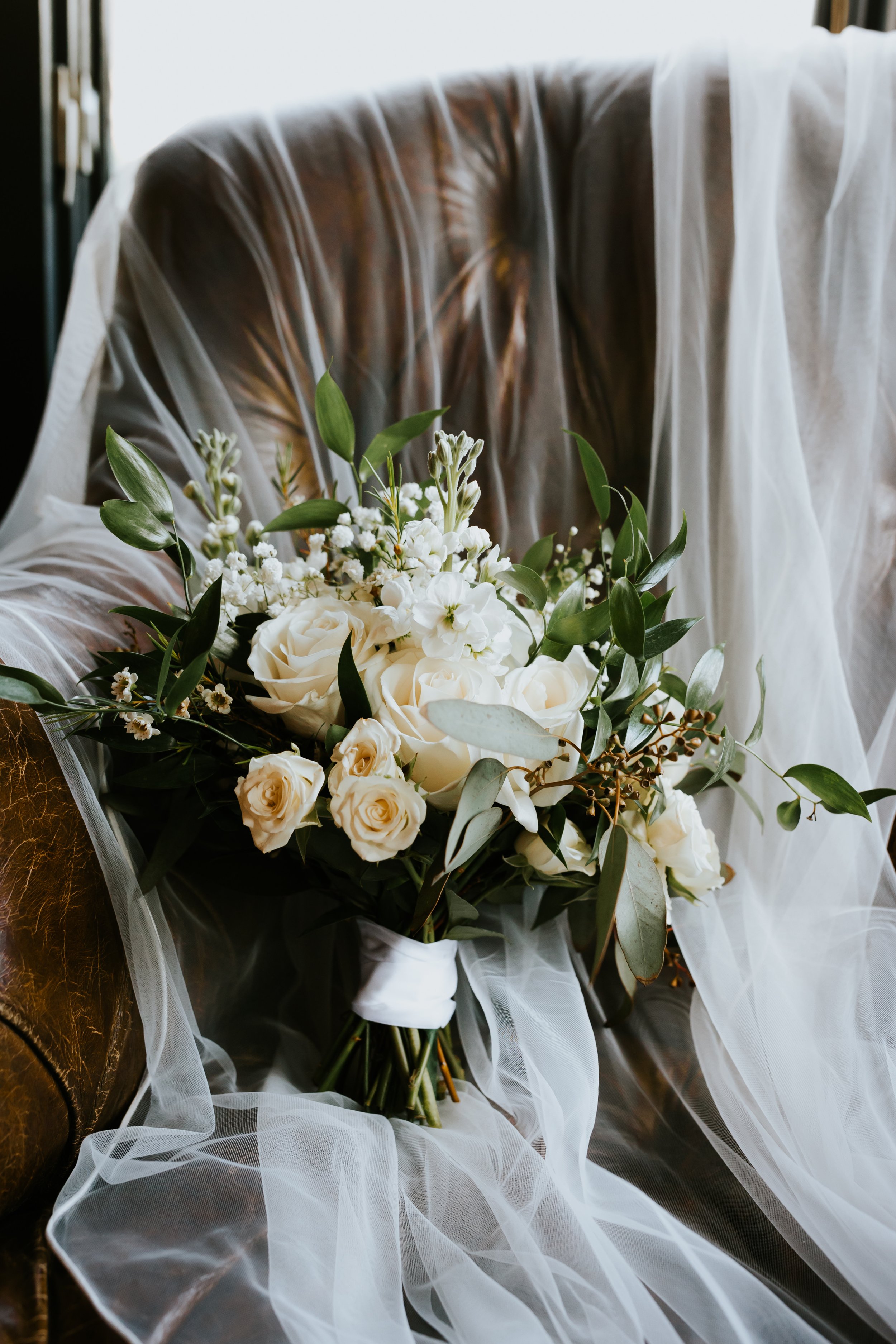 A bouquet of white roses, baby's breath, and greenery, resting on a sheer white fabric, with a dark background.