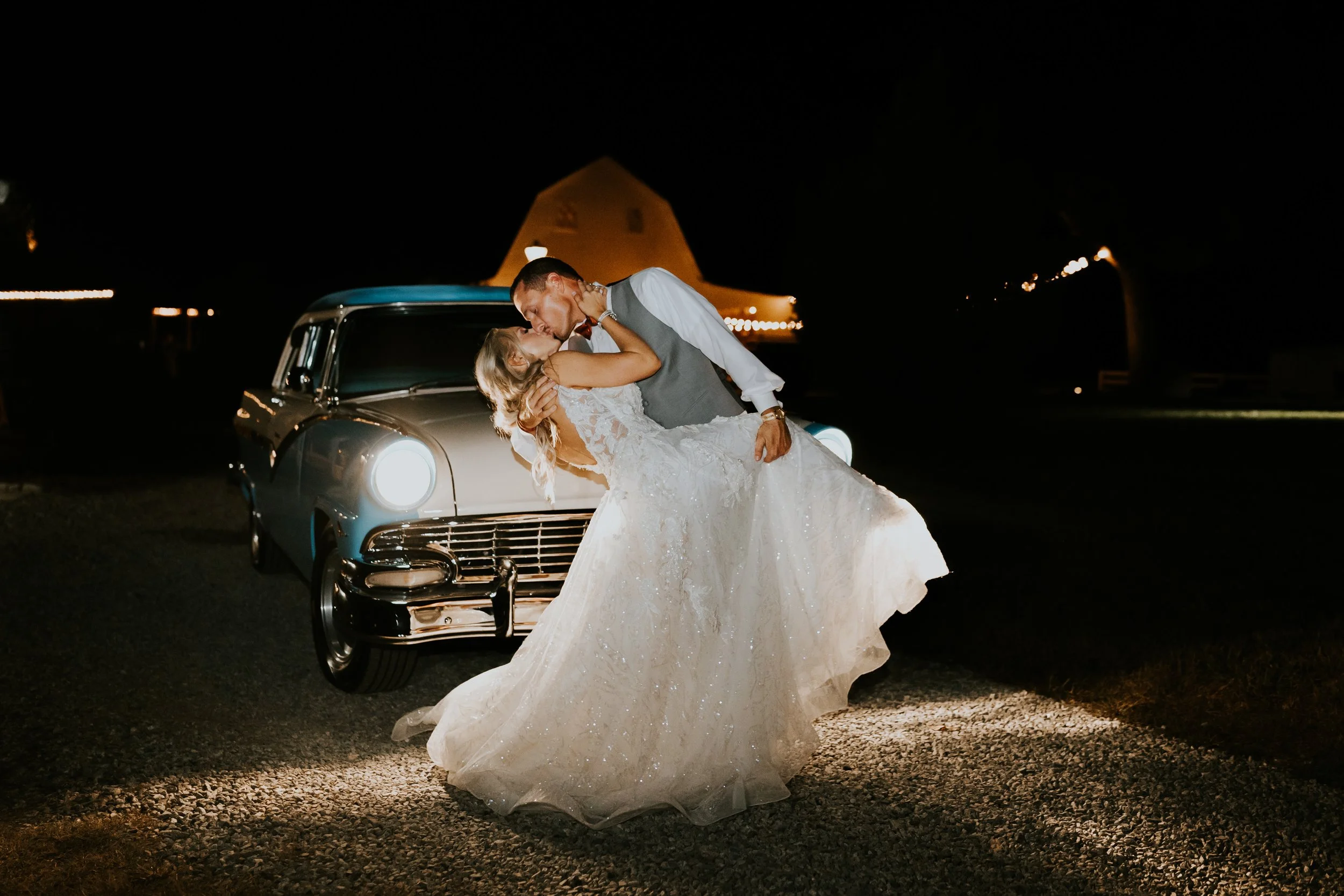 A newlywed couple sharing a kiss at night, with the groom carrying the bride in front of a vintage car and a barn with string lights in the background.