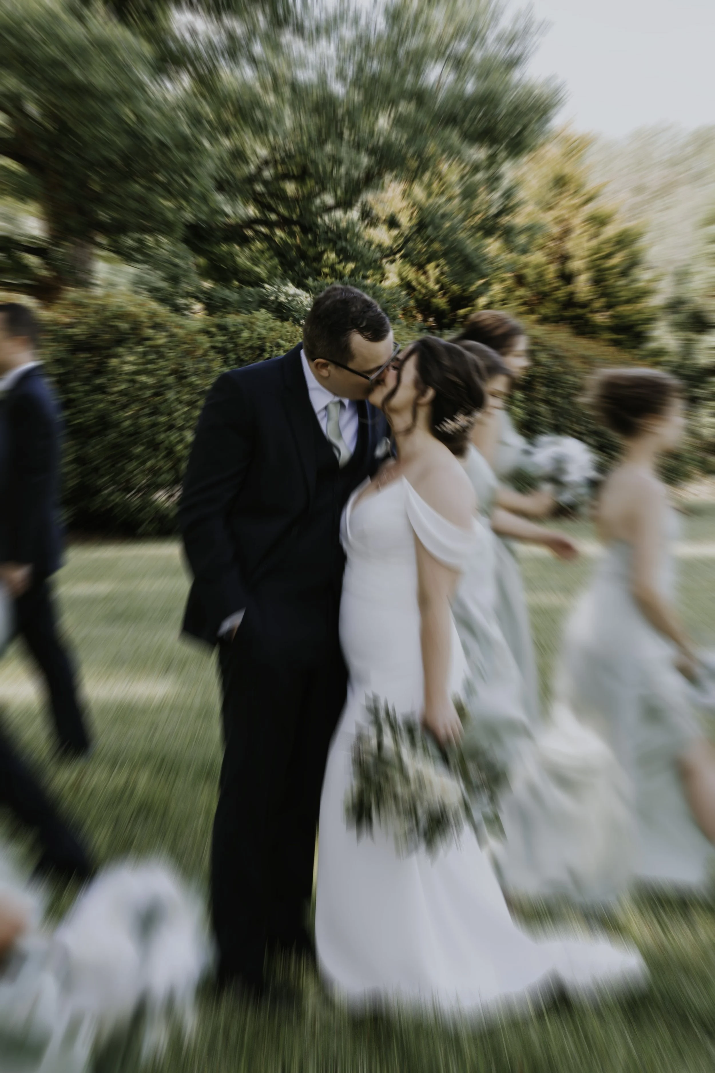 A bride and groom sharing a kiss at an outdoor wedding with trees in the background, with a motion blur effect.