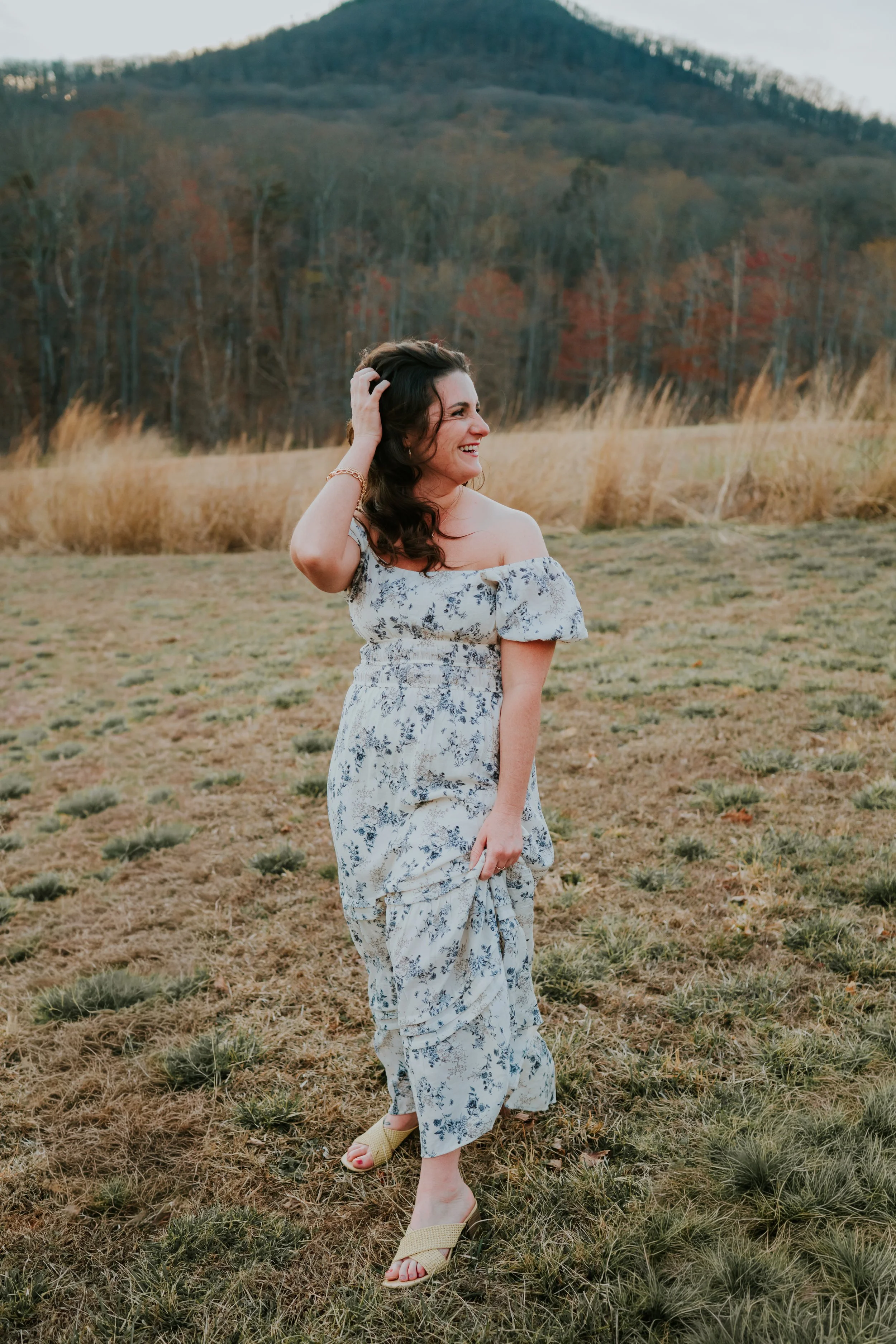 A woman in a floral off-shoulder dress standing in a field with a mountain in the background, smiling and touching her hair.