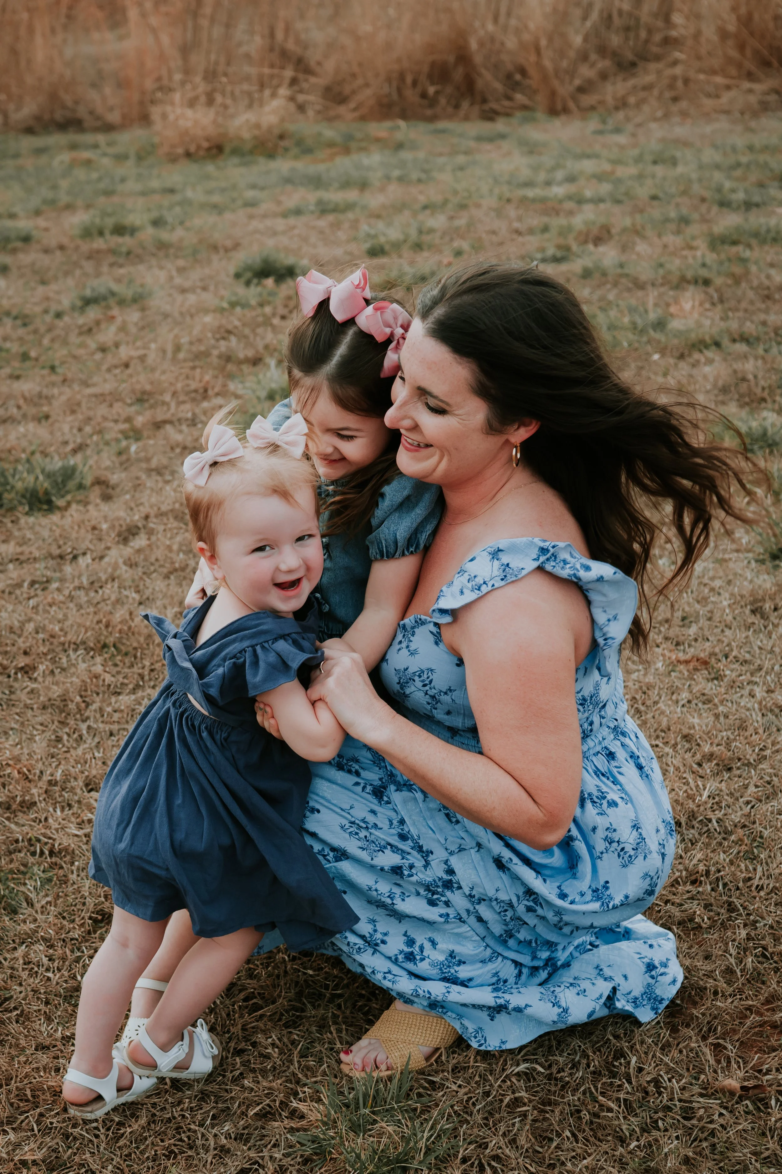 A woman and two young girls embracing and smiling outdoors on a grassy field.