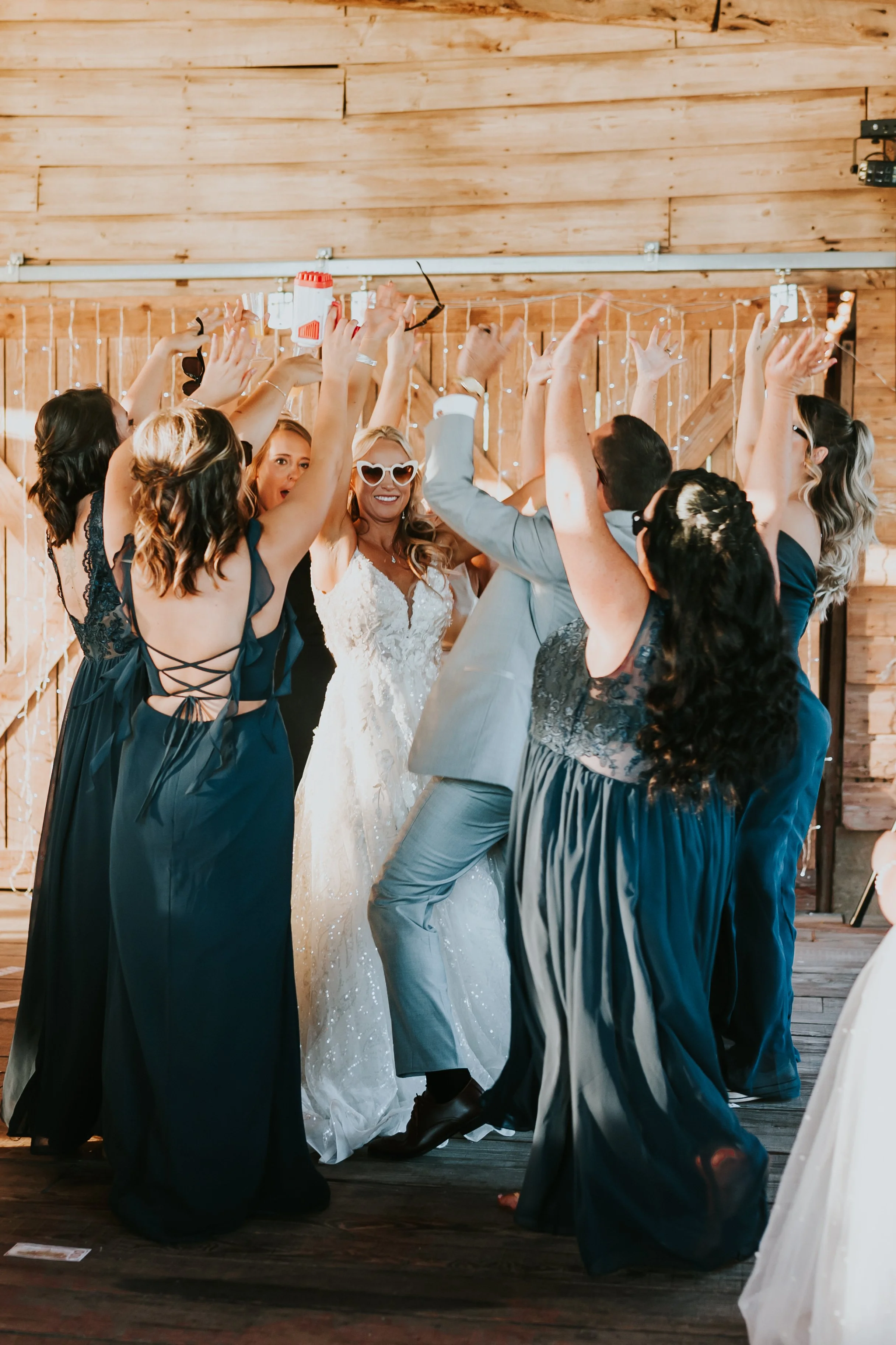 A group of people at a wedding celebration dancing together, with the bride wearing sunglasses and a white wedding dress, and others in formal attire, in a rustic wooden venue.