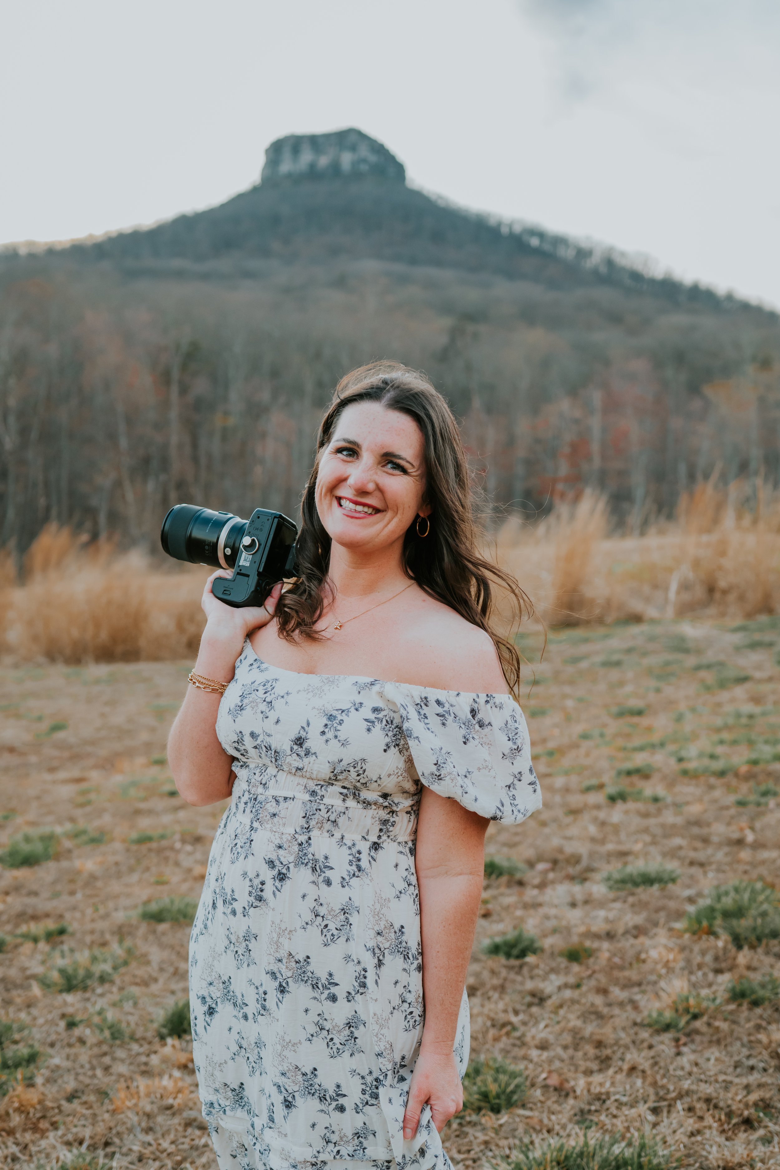 A woman with long brown hair smiling outdoors, holding a camera on her shoulder, with a mountain and trees in the background.
