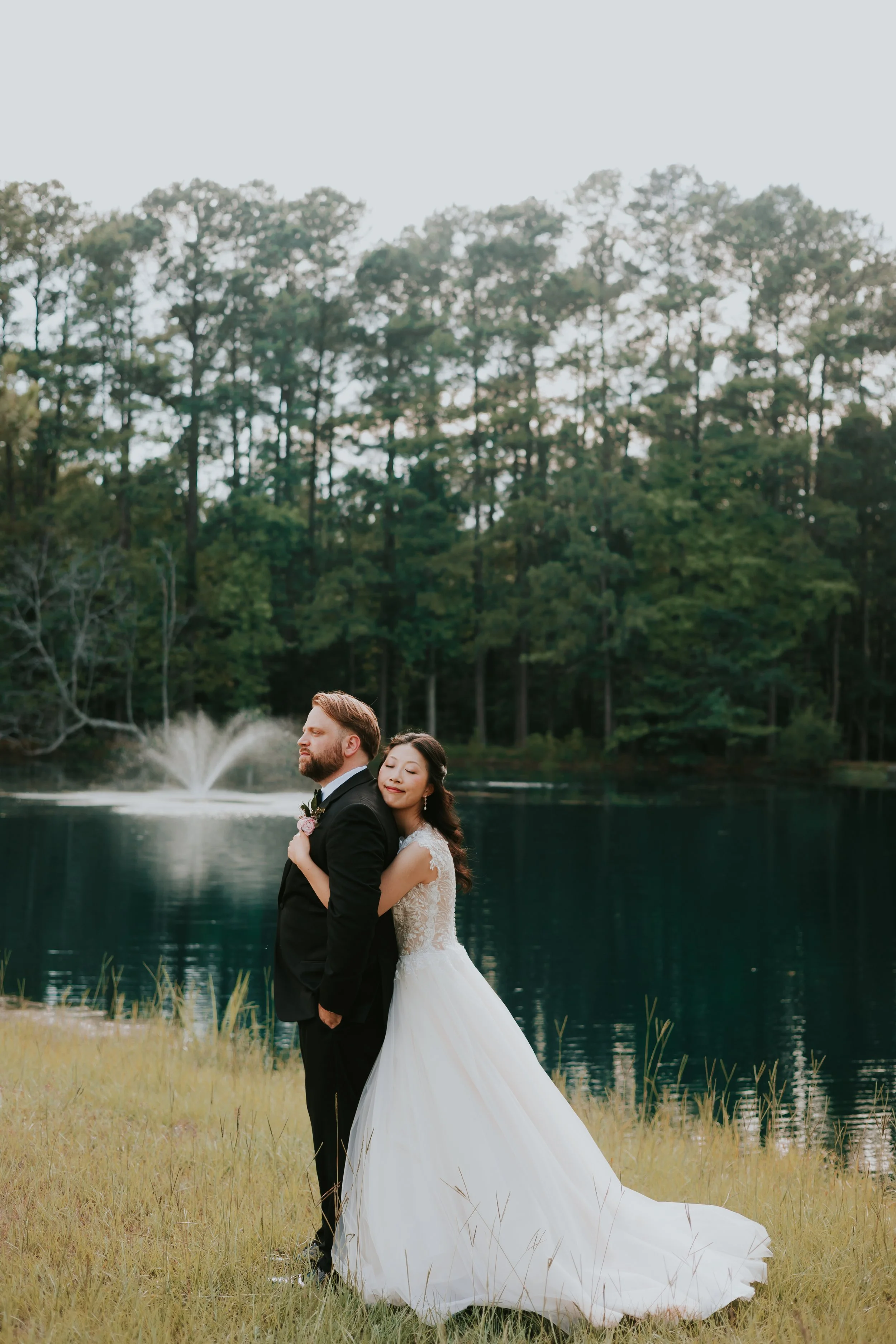 A bride and groom standing by a lake with a fountain in the background, surrounded by trees, on a grassy bank.