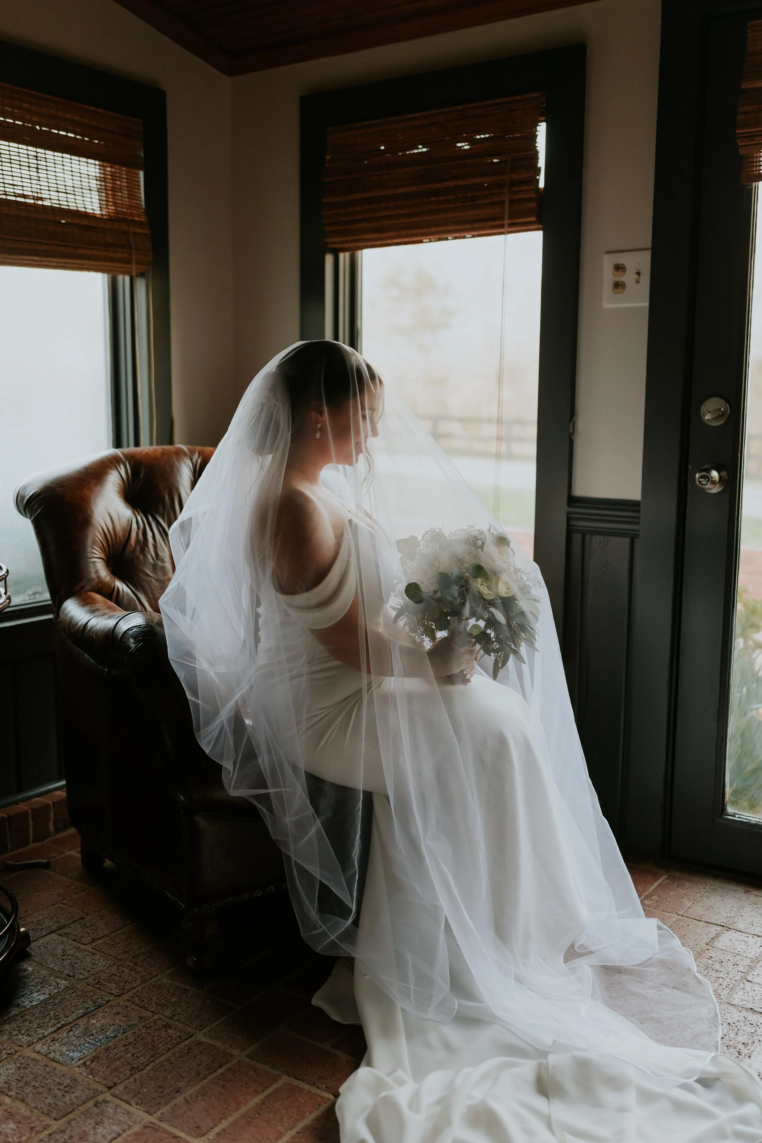 Bride sitting on a leather armchair, wearing a wedding dress and veil, holding a bouquet of flowers, indoors by a window with wooden blinds.
