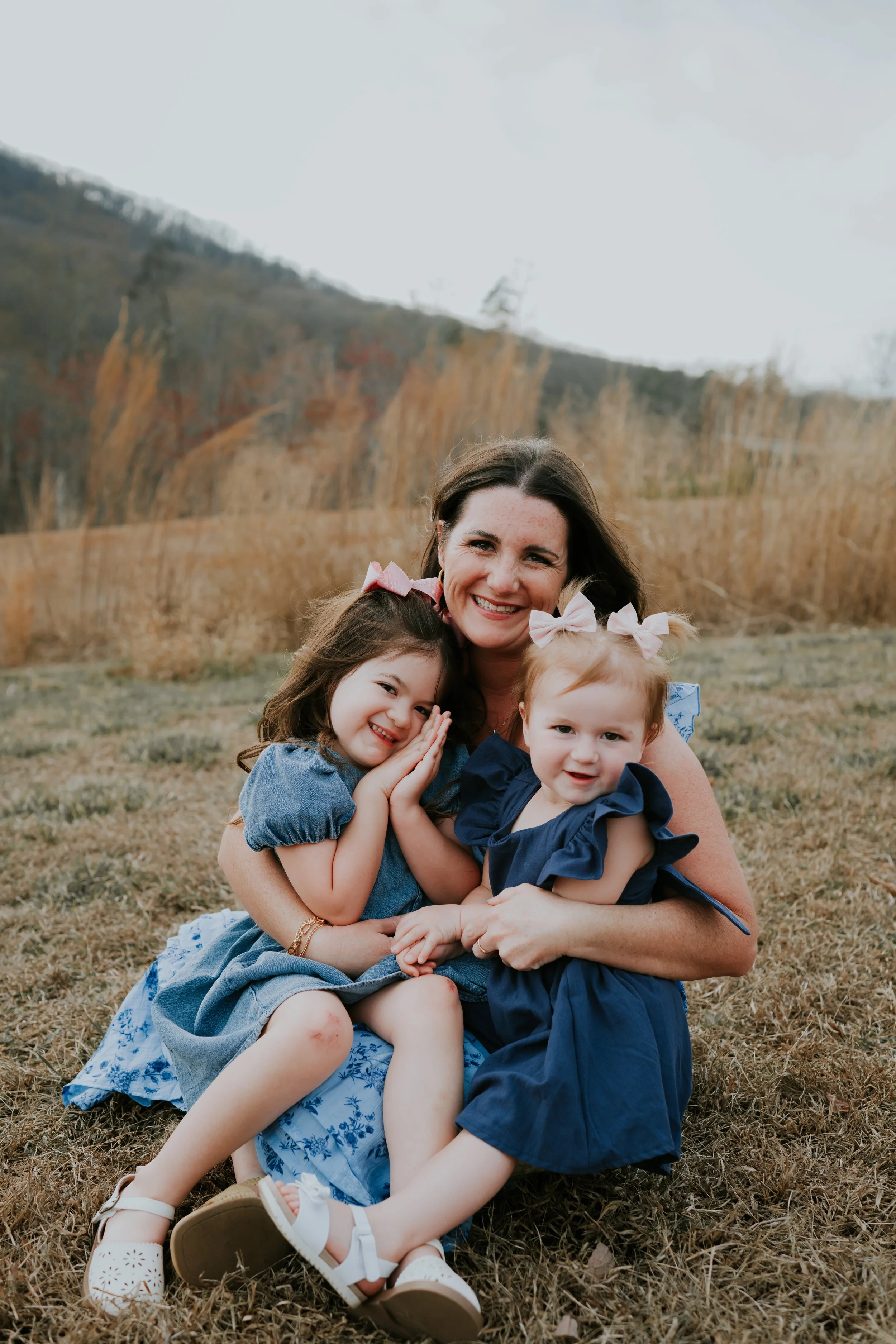 A woman with long brown hair smiling and sitting on the grass in a field, hugging three young girls, all wearing blue dresses with pink or blue bows in their hair, with a hillside and dry grass in the background.