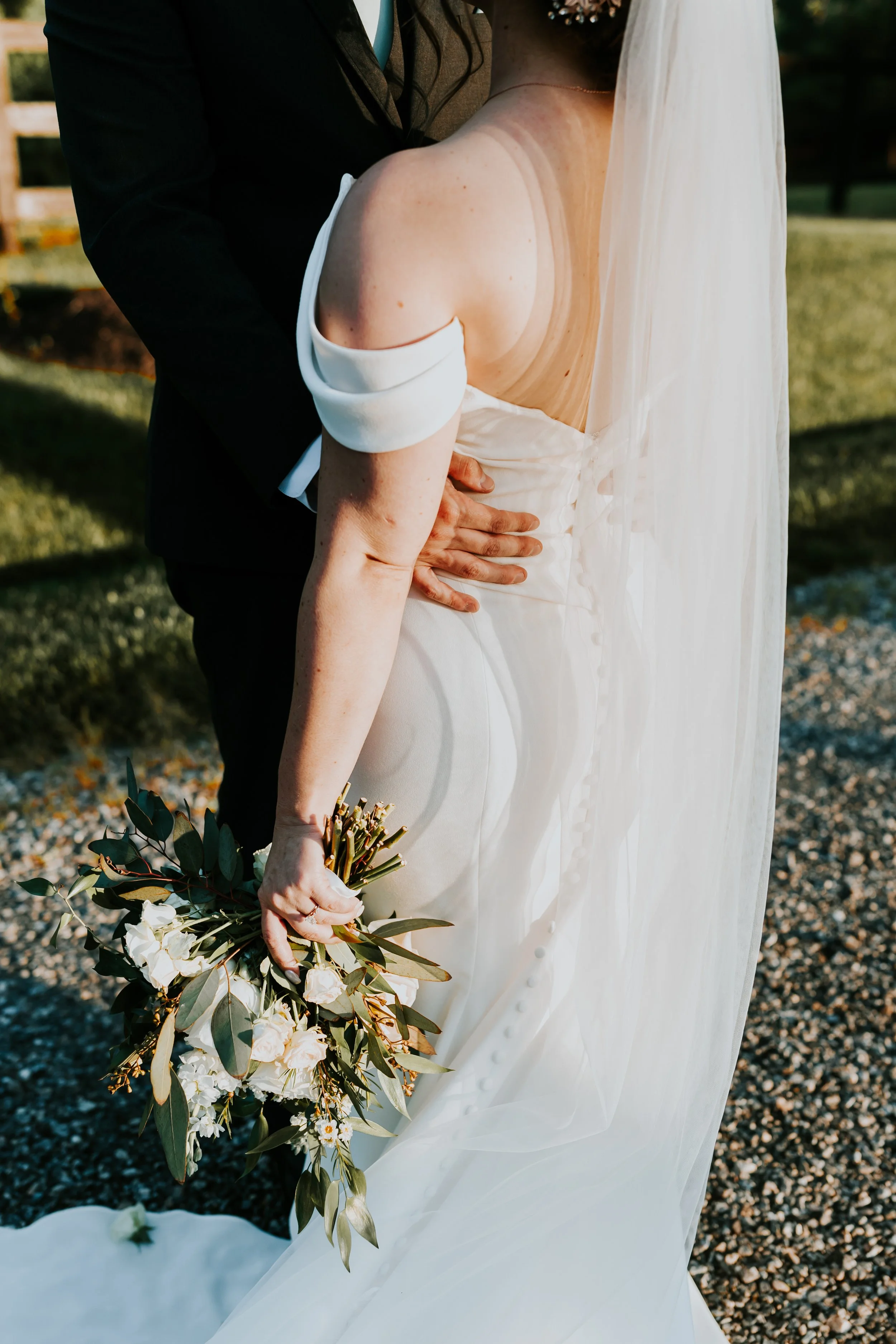 A bride holding a bouquet of white and green flowers, wearing a white off-shoulder wedding dress with buttons down the back, in an outdoor setting.