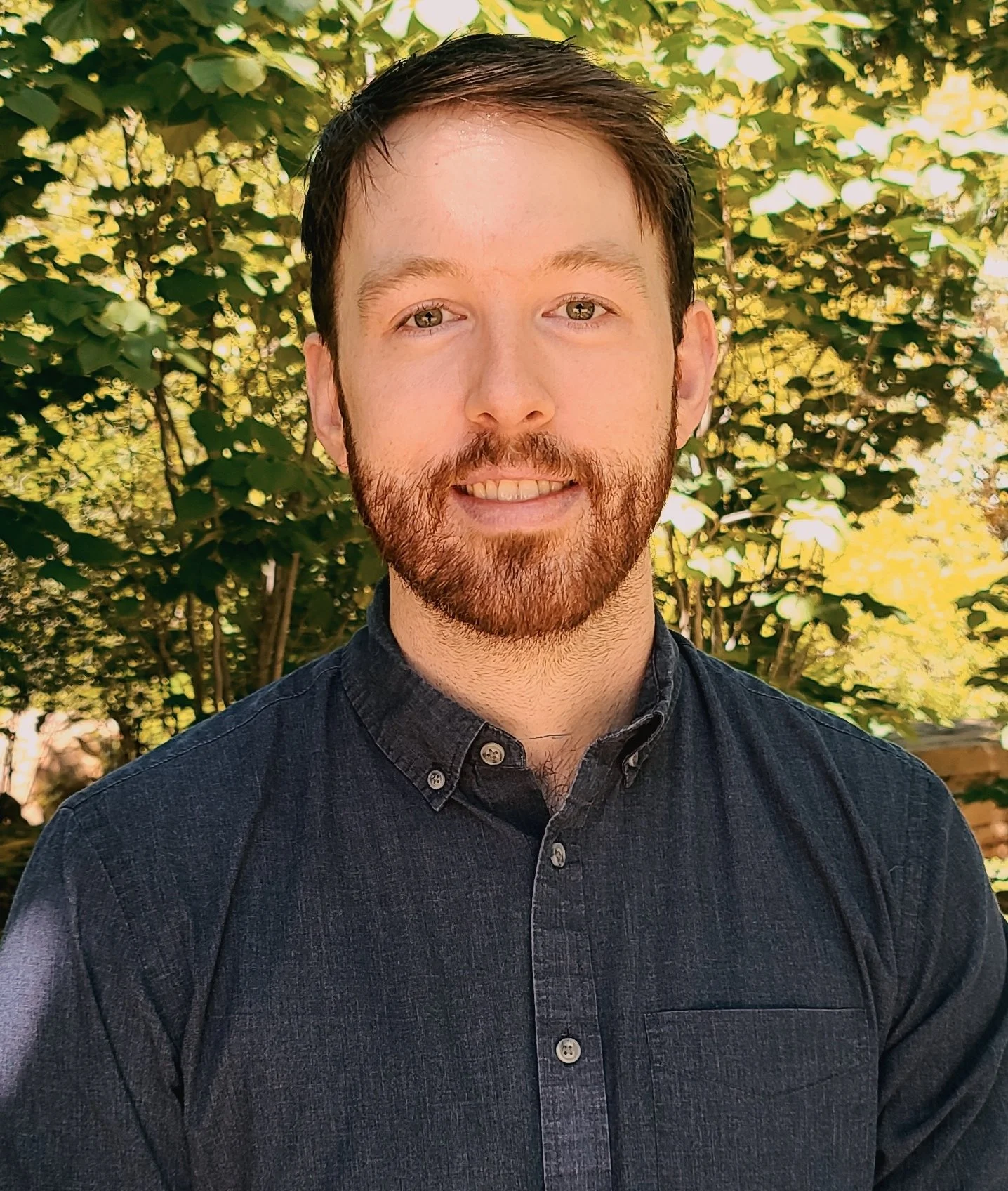 A young man with brown hair and a beard smiling outdoors in front of green leafy trees, wearing a dark button-up shirt.