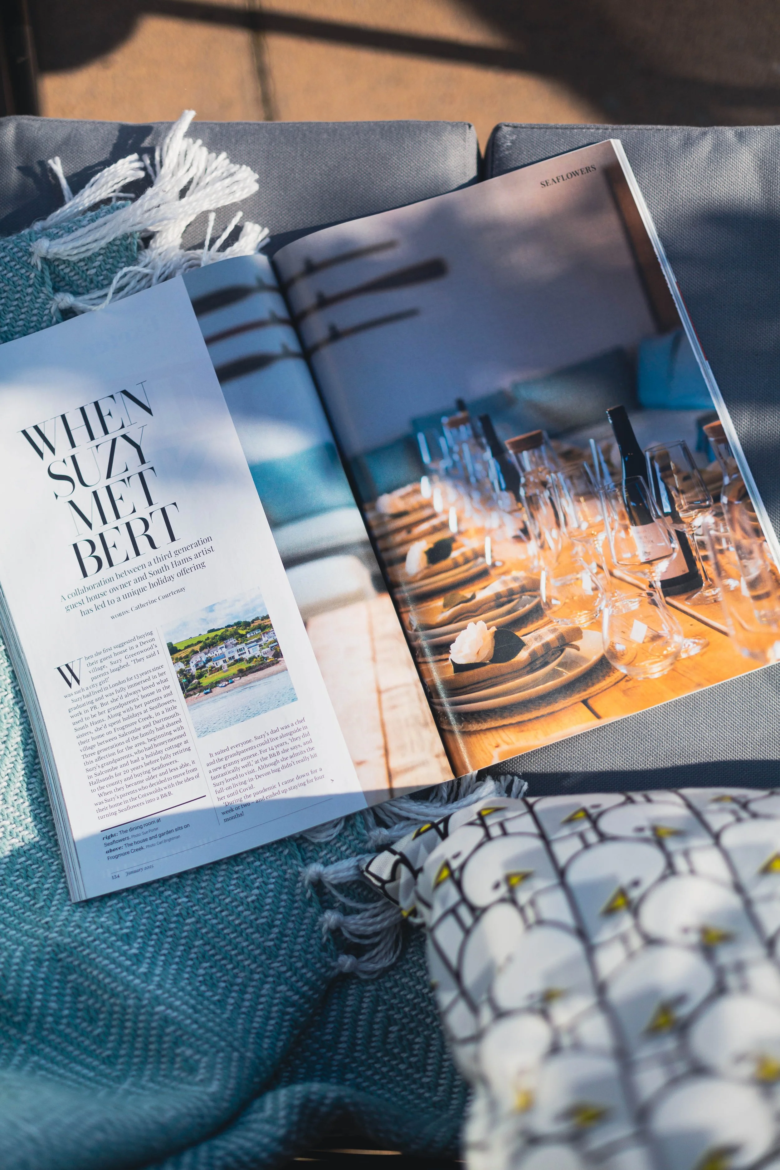 Open magazine on a blue woven surface, with a woven scarf and a patterned cloth nearby, sunlight illuminating the scene.