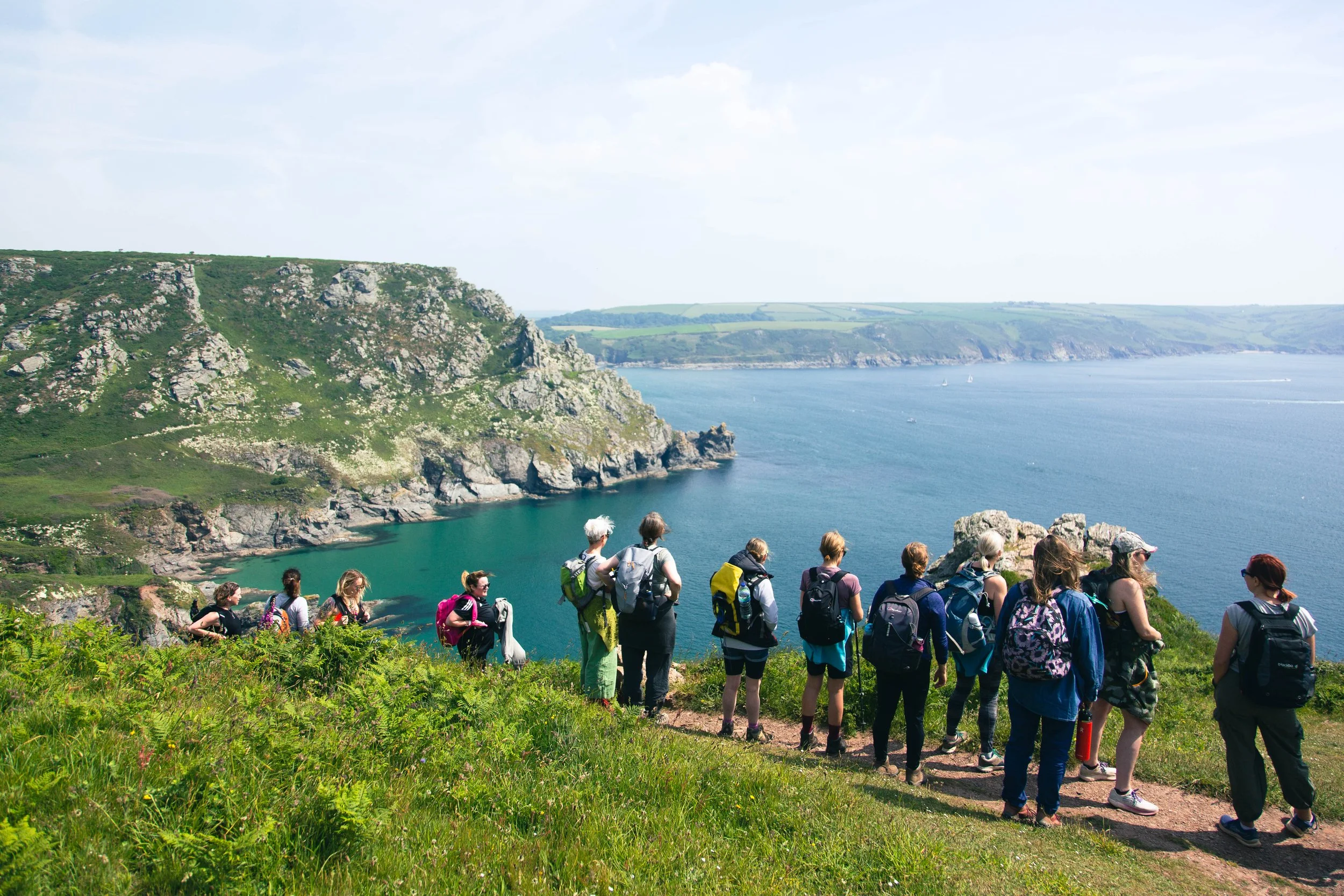 Group of hikers standing on a grassy hill overlooking a coastal scene with green cliffs, a cove with blue water, and sailboats in the distance.