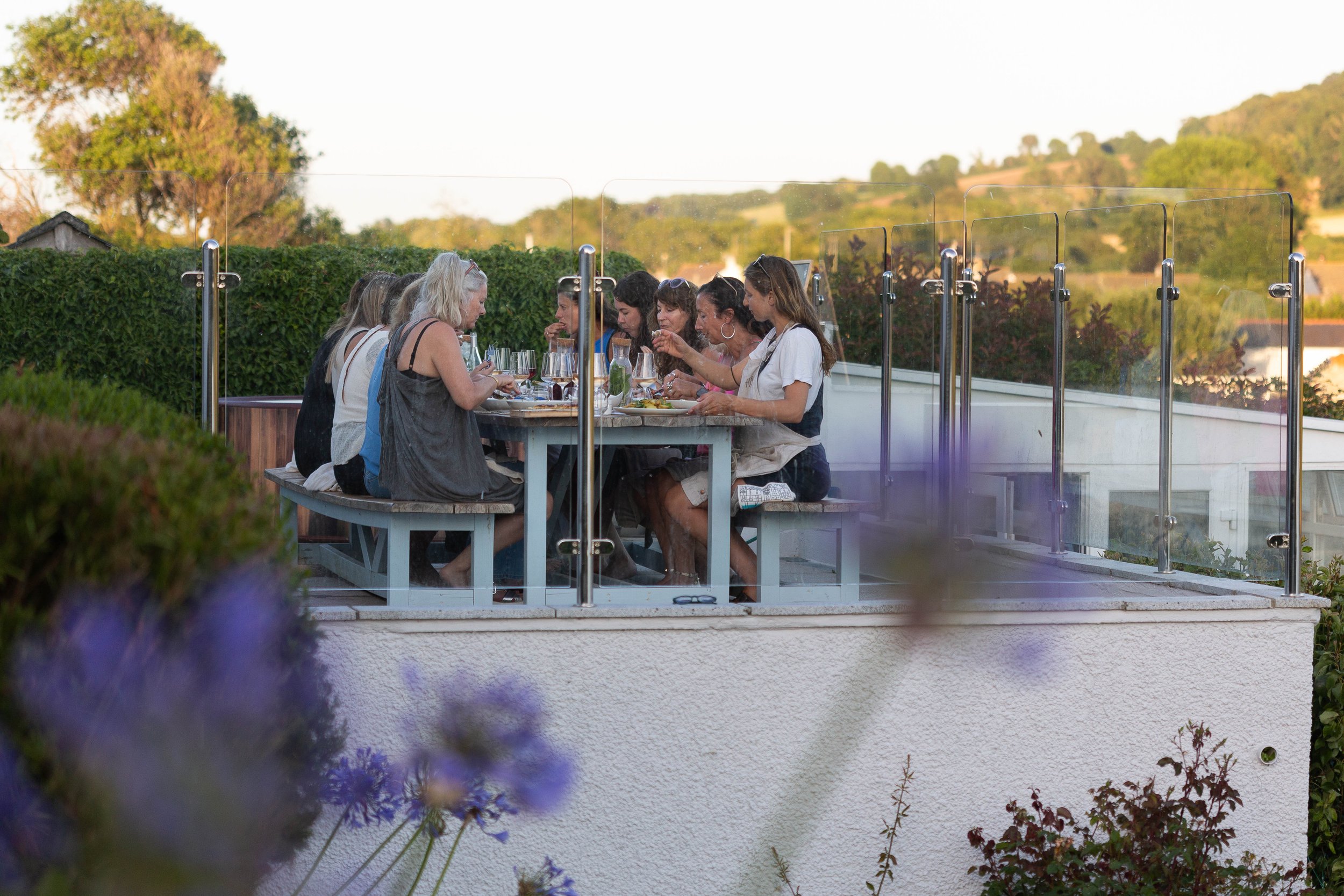Group of women sitting around a table outside on a patio, enjoying a meal with glasses of wine, with a green hedge, trees, and hills in the background at sunset.