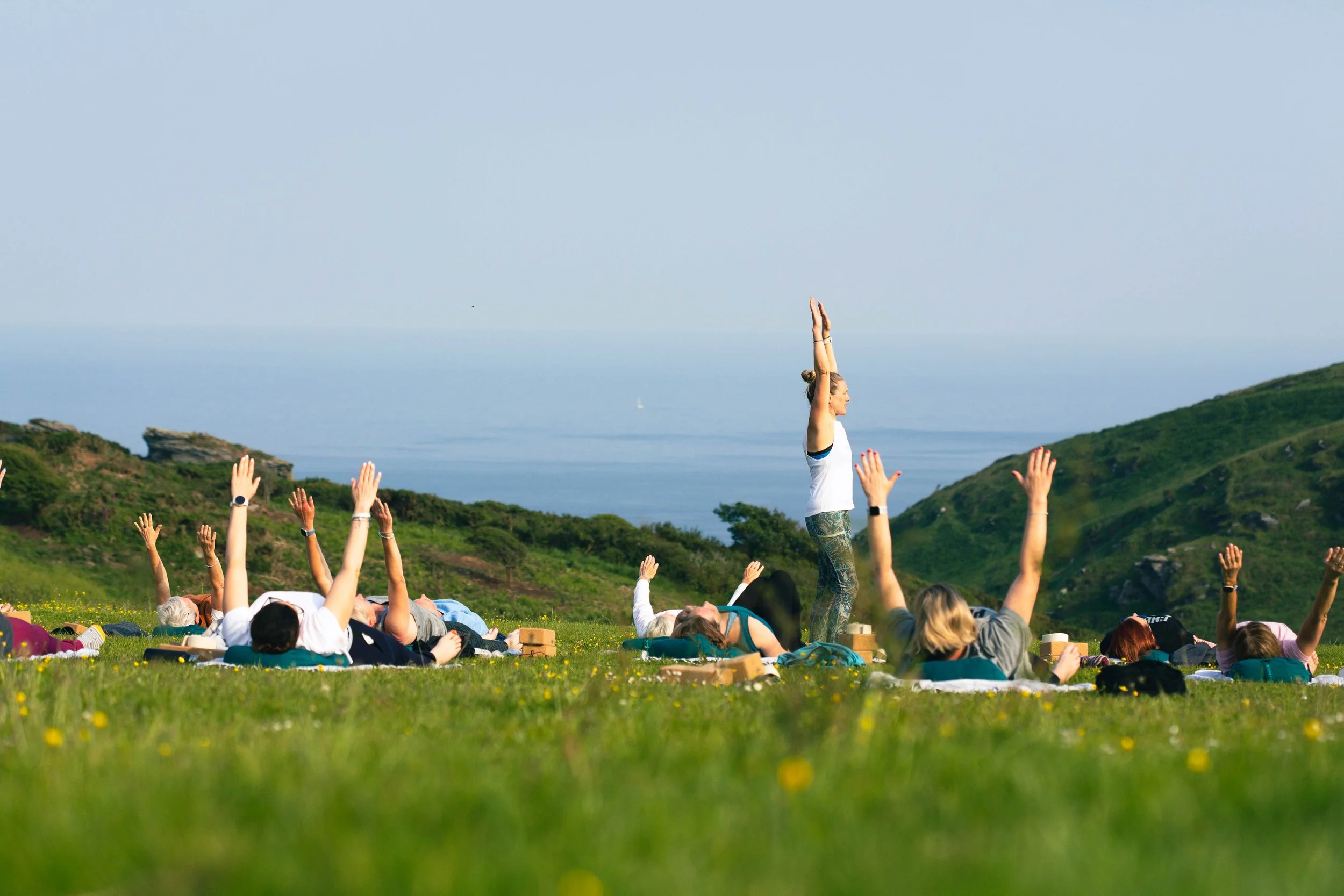 A group of people practicing yoga outdoors on grass near a coastline, with trees and hills in the background and a person standing in a yoga pose while others lie on mats.