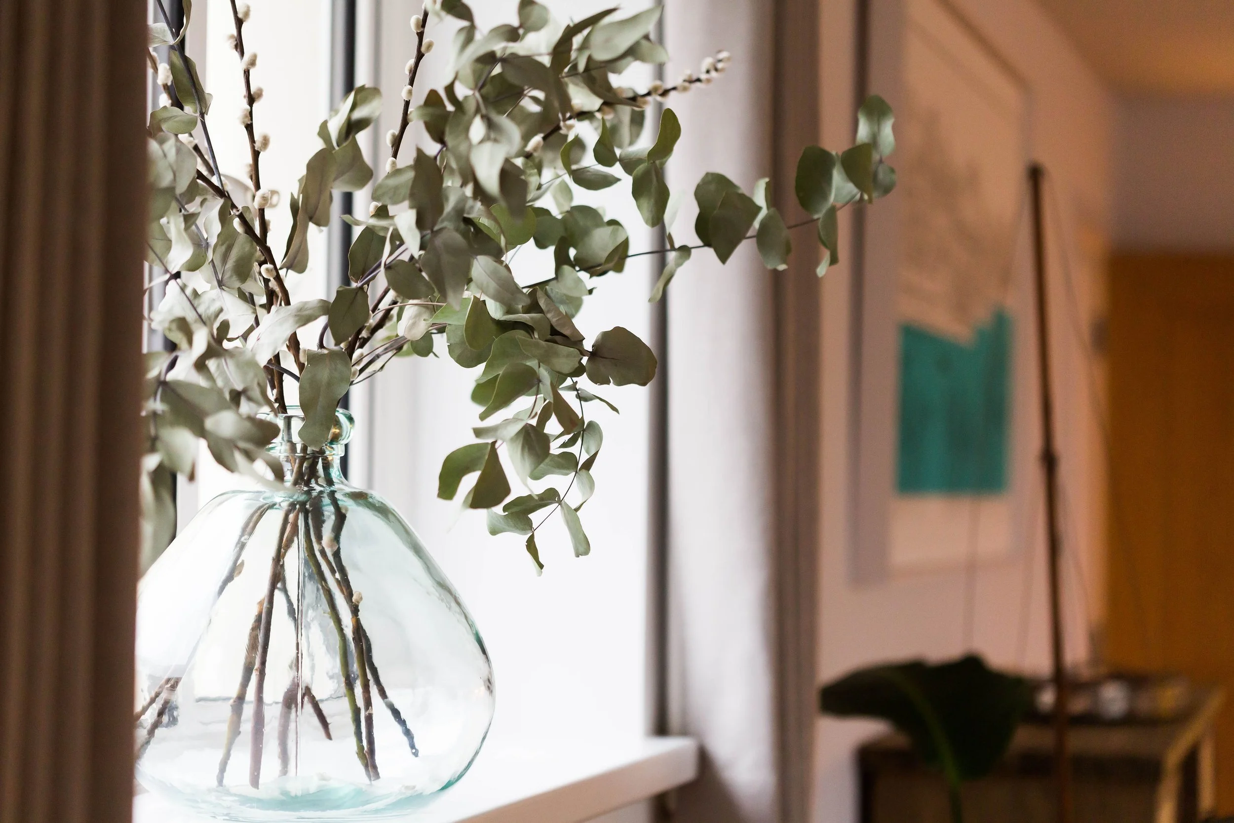A glass vase with green leafy stems placed on a window sill, with natural light coming through the window.