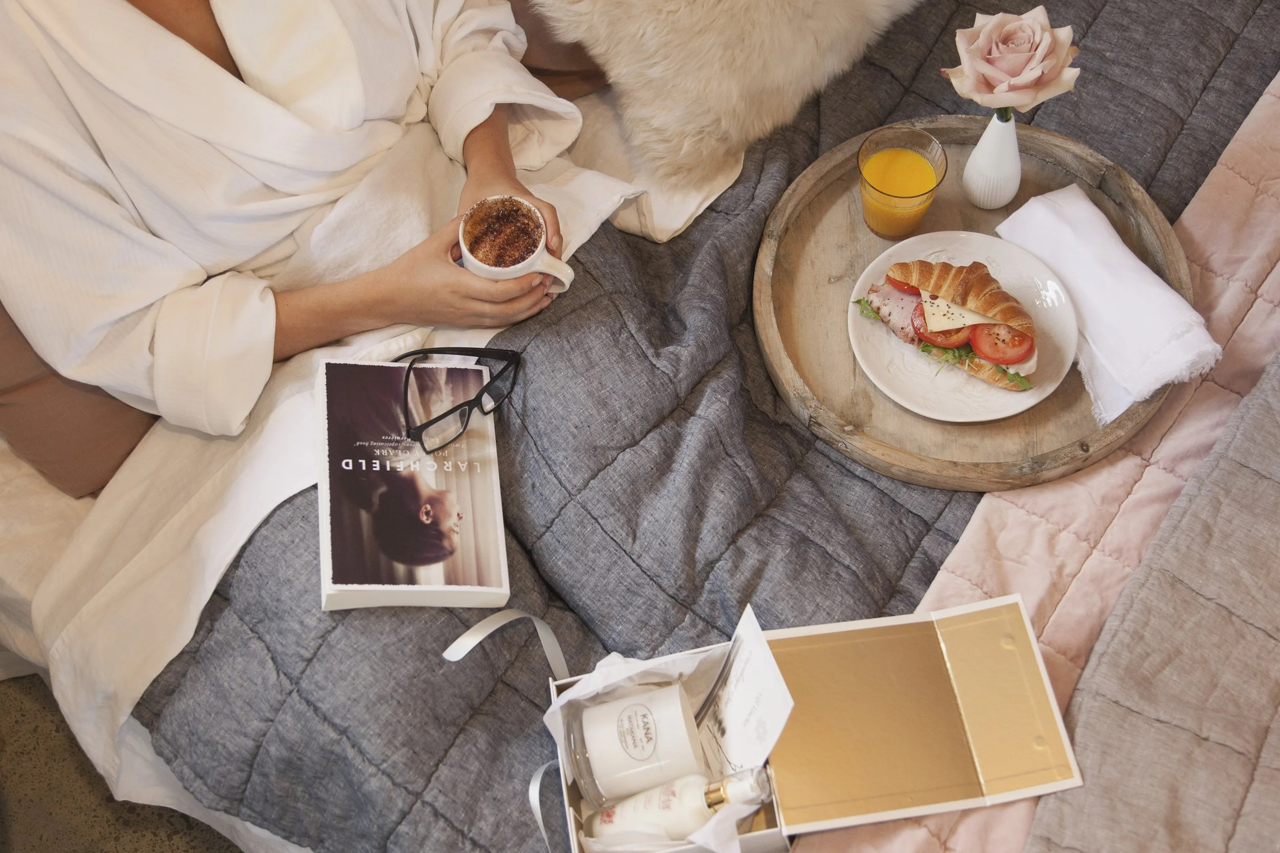 A woman in a white robe sitting on a gray bed, holding a mug of coffee topped with cinnamon. On the bed are a pair of glasses, a magazine, and a small white bag. Next to the bed, a wooden tray with a croissant sandwich, a glass of orange juice, and a white vase with pink roses. Also visible are a tissue box and a brown and gold box, all arranged on the bed and side table.