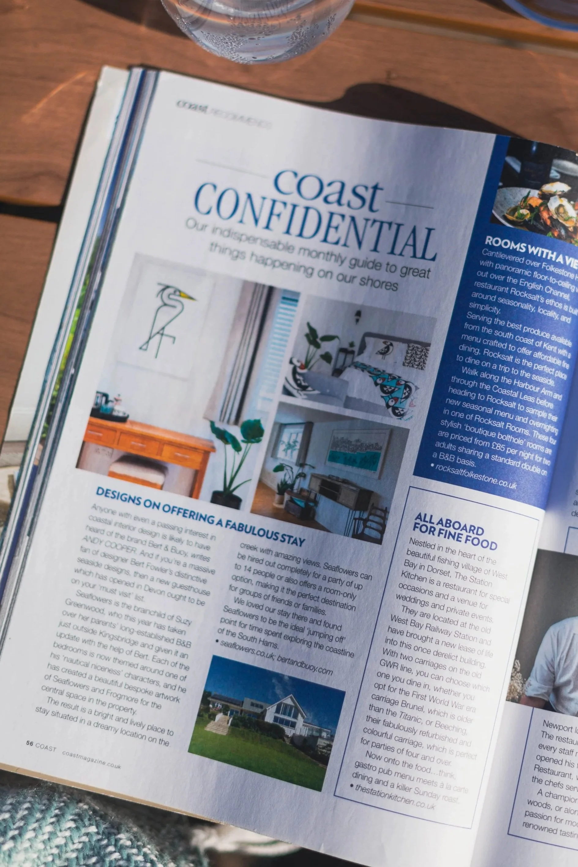 Open magazine page showing a guide titled 'Coast Confidential' with images of interior decor and a house exterior, placed on a wooden surface with a glass of water nearby.