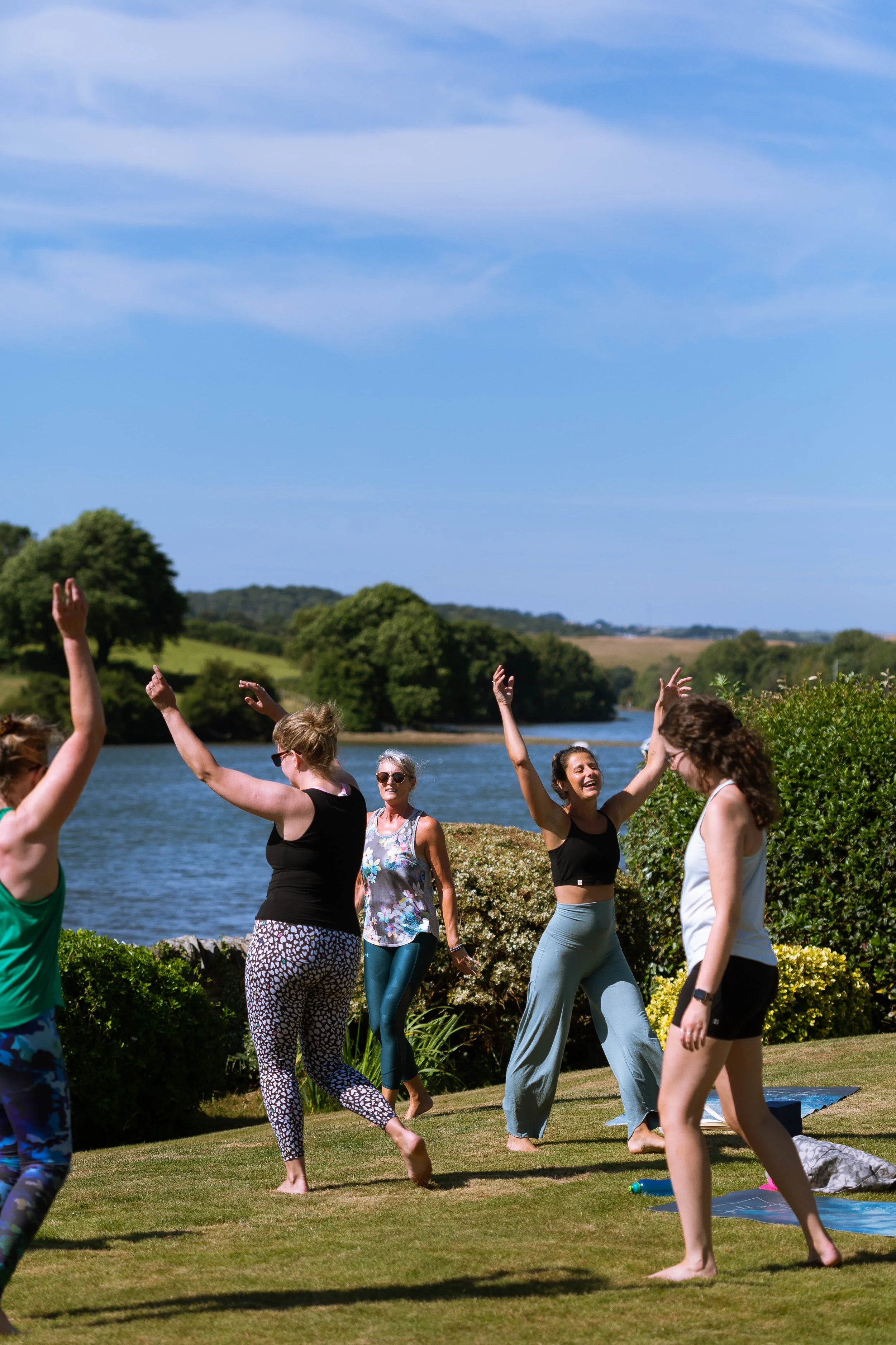 People practicing yoga outdoors near a lake, with trees and a blue sky in the background.