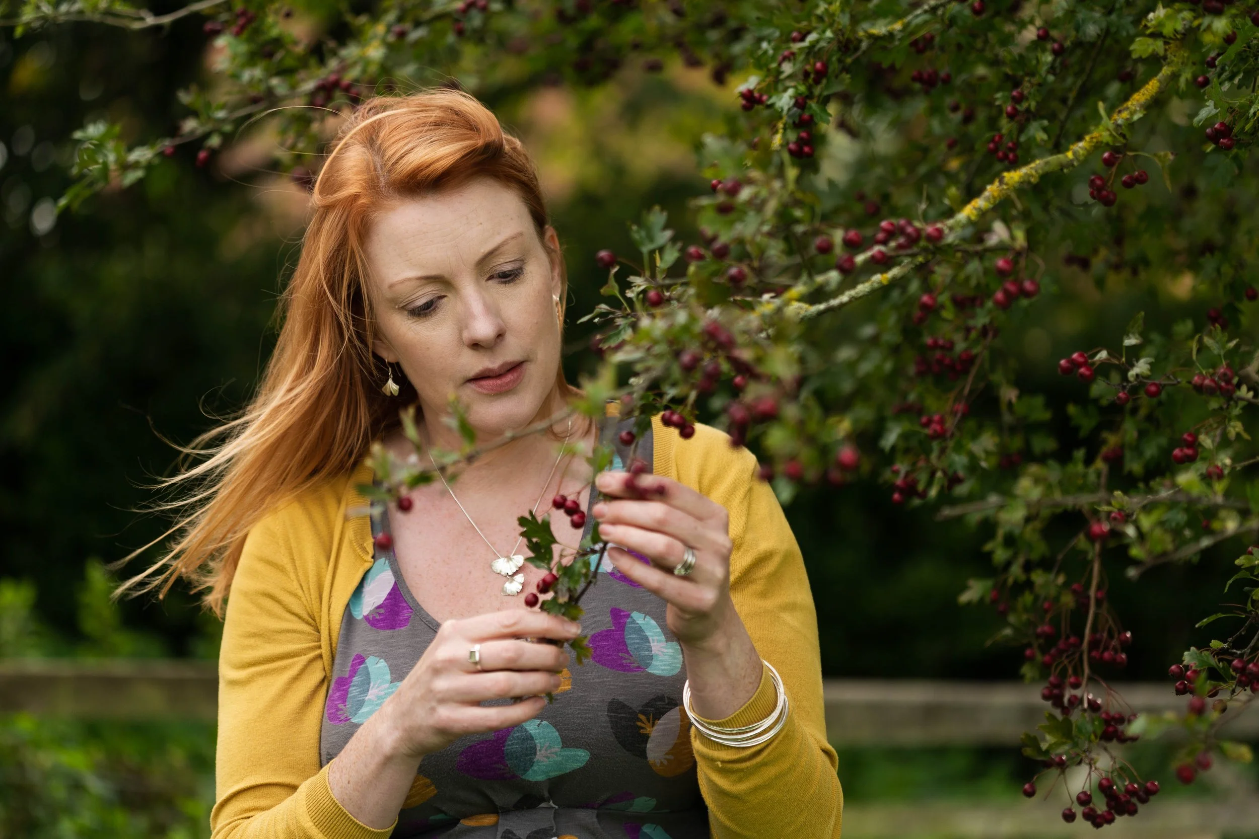 A woman with red hair and jewelry examining red berries on a bush in a lush outdoor setting.
