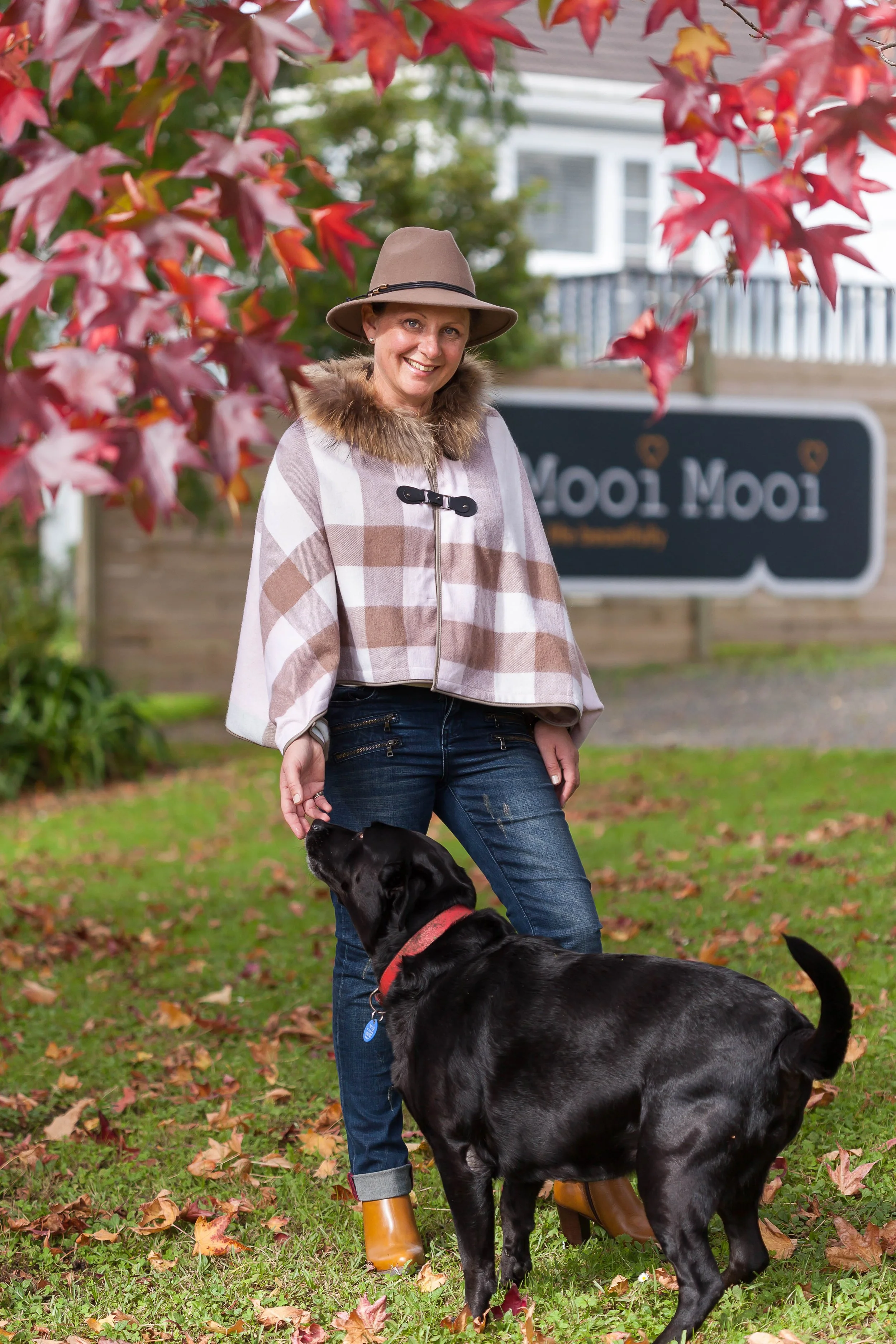 A woman smiling outdoors, wearing a beige hat, a beige and white checkered coat with a fur collar, blue jeans with rolled-up cuffs, and yellow rain boots, stands on a grassy area with fallen autumn leaves, next to a black Labrador retriever dog. In the background, there is a wooden fence and a sign that says "Moo! Moo!" with a blurred residential house behind it, and red and green autumn foliage surrounds the scene.