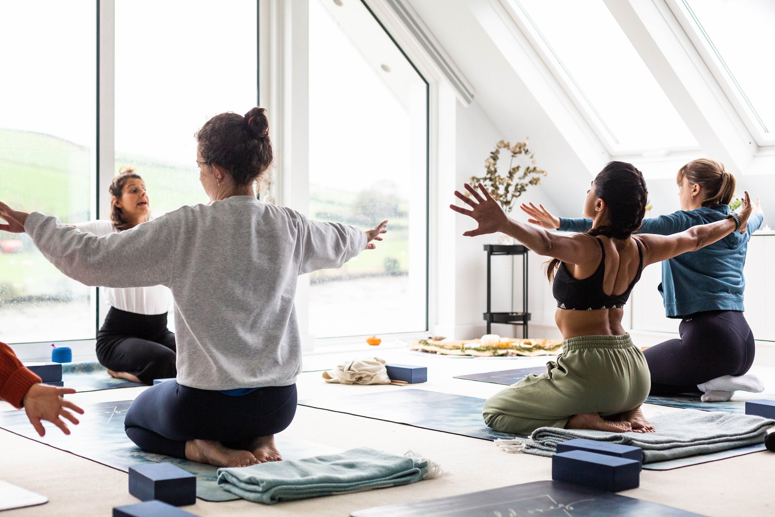 Group of people practicing yoga or meditation together in a bright room with large windows, sitting cross-legged on mats with arms extended outward.