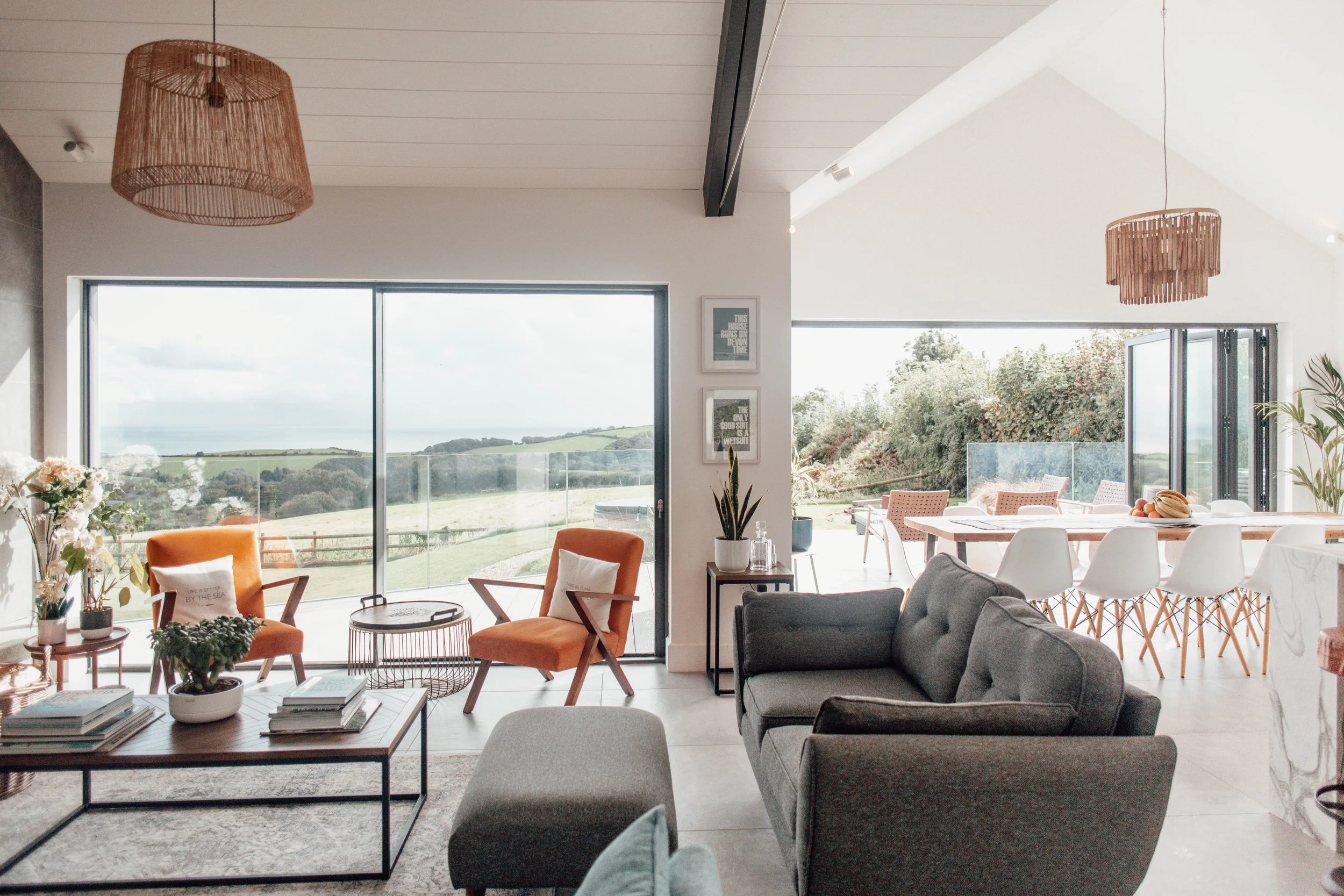 Open living room with large windows overlooking a green landscape, featuring a gray sofa, orange chairs, a coffee table, and a dining area with white chairs and a wooden table.