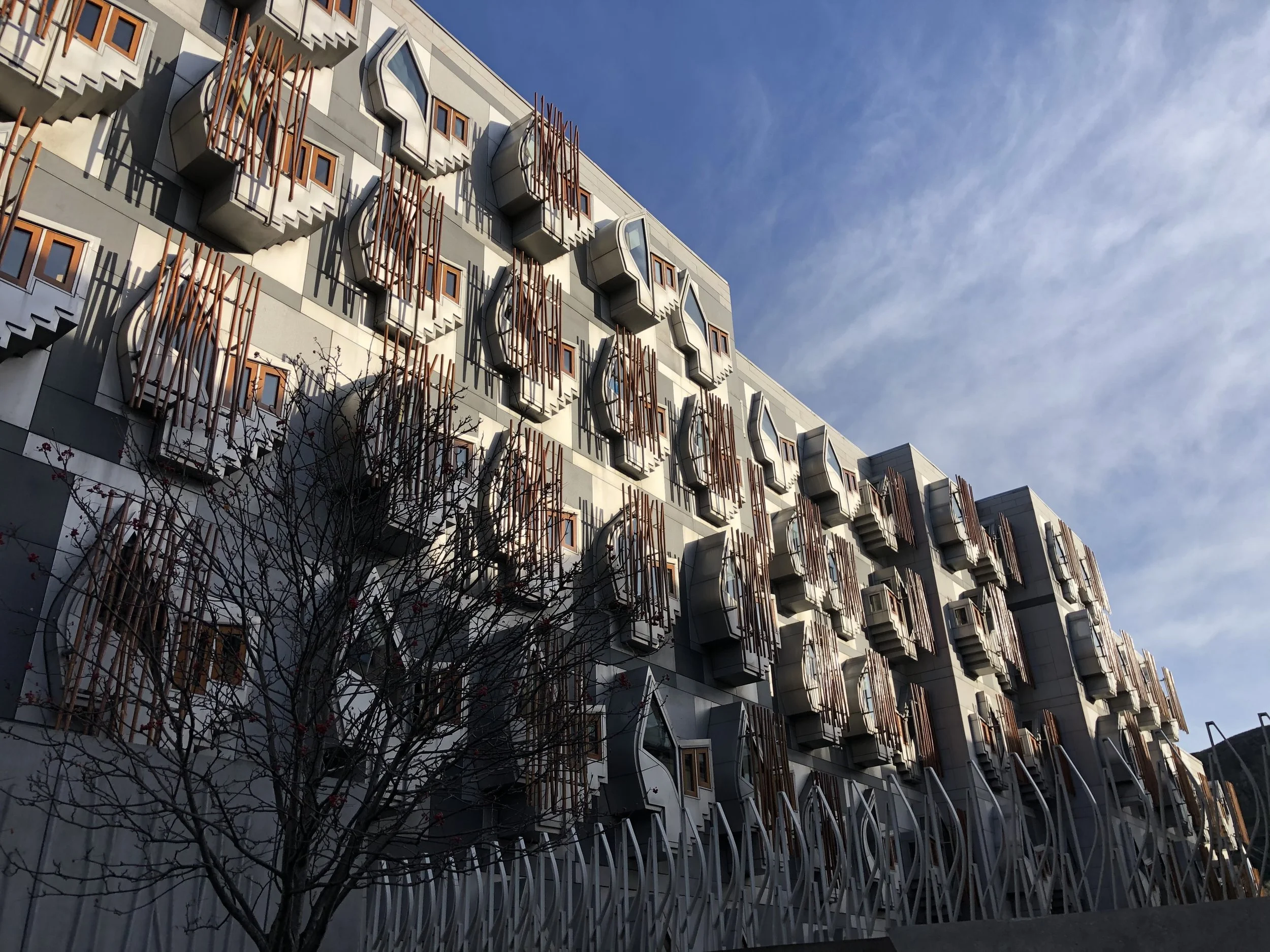 A modern building with decorative protruding balconies and windows, along with orange metal railings. The sky is mostly clear with some streaked clouds.