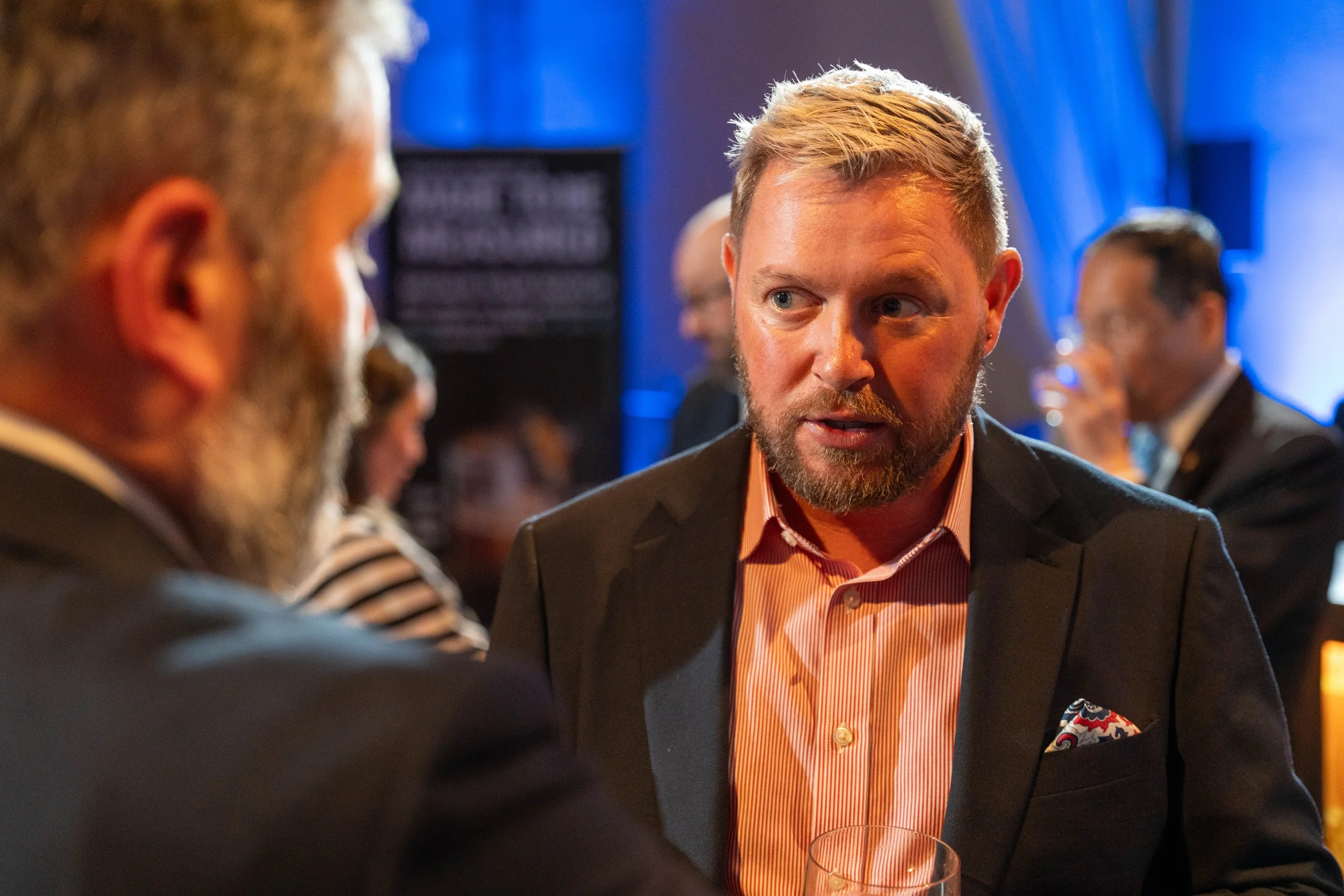 A man with blond hair and a beard wearing a dark suit jacket and a light orange striped shirt, engaged in conversation at a social event, with blurred people and blue lighting in the background.