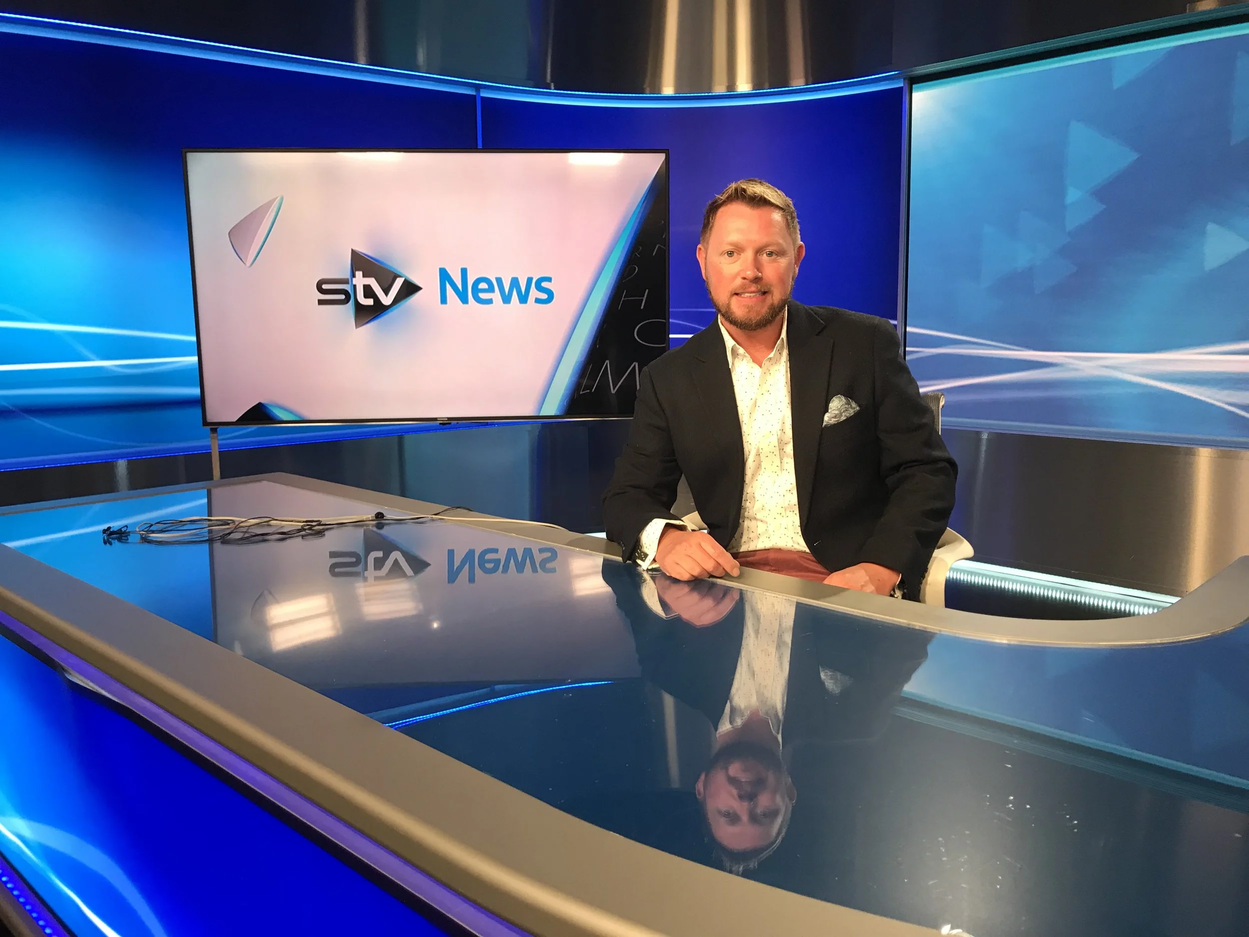 A man in a black suit and white shirt sitting behind a news desk, smiling. Behind him is a large screen displaying the logo for STV News and a blue background with light streaks.