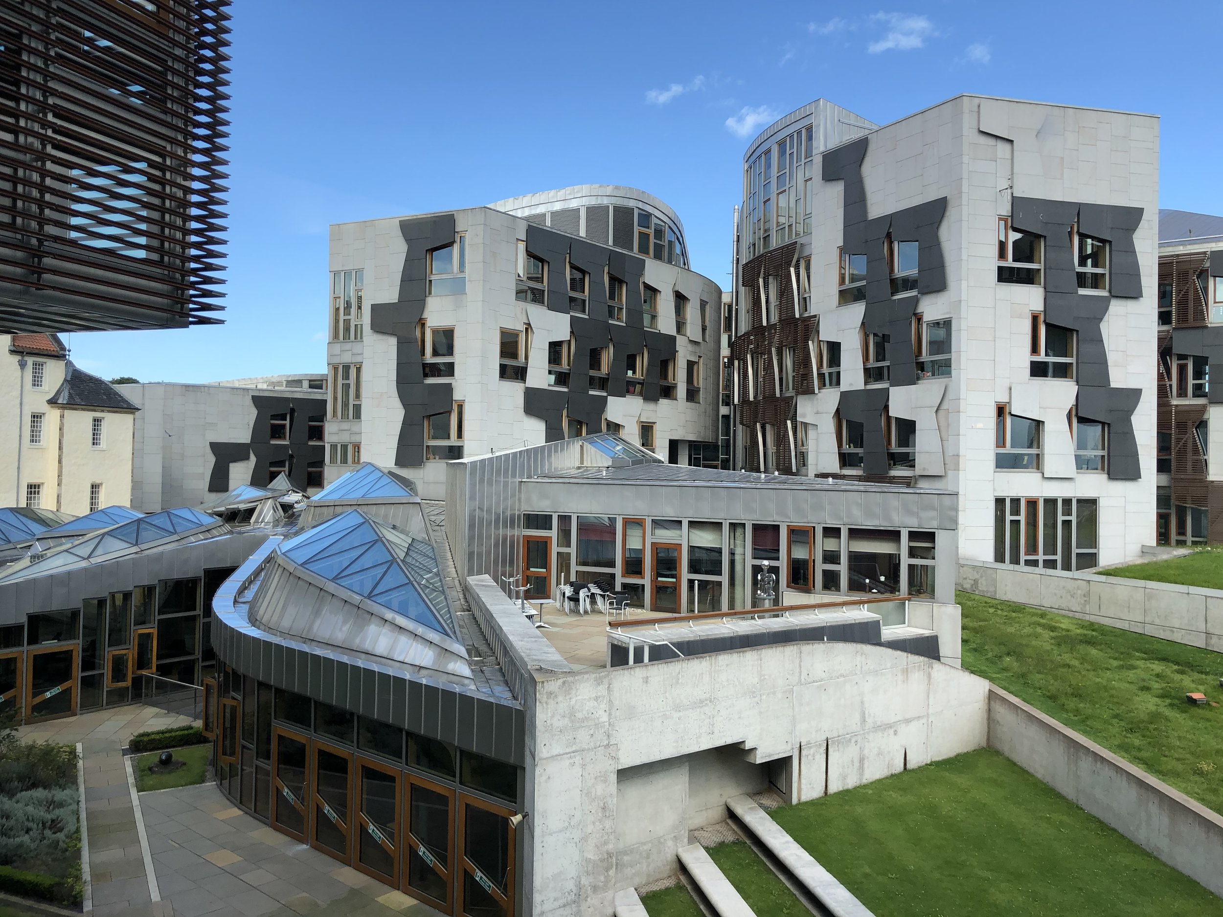 Modern building with curvy, white and gray exterior, numerous windows, and rooftop structures, surrounded by green lawn and adjacent older buildings, under a clear blue sky.