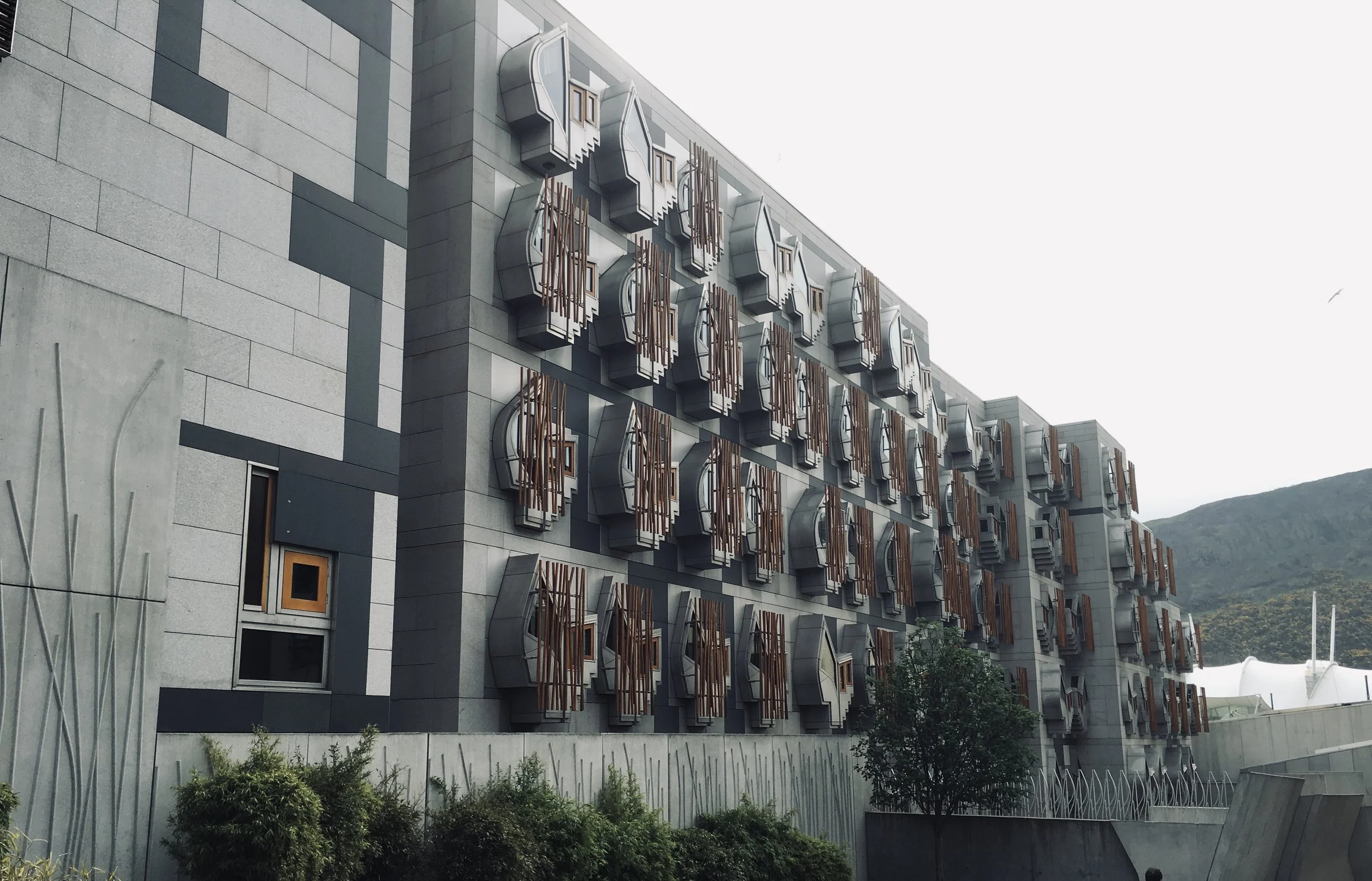 Modern building with unique balcony design featuring protruding, rounded structures with wooden accents, over a landscaped area with small bushes and trees, mountains in the background.