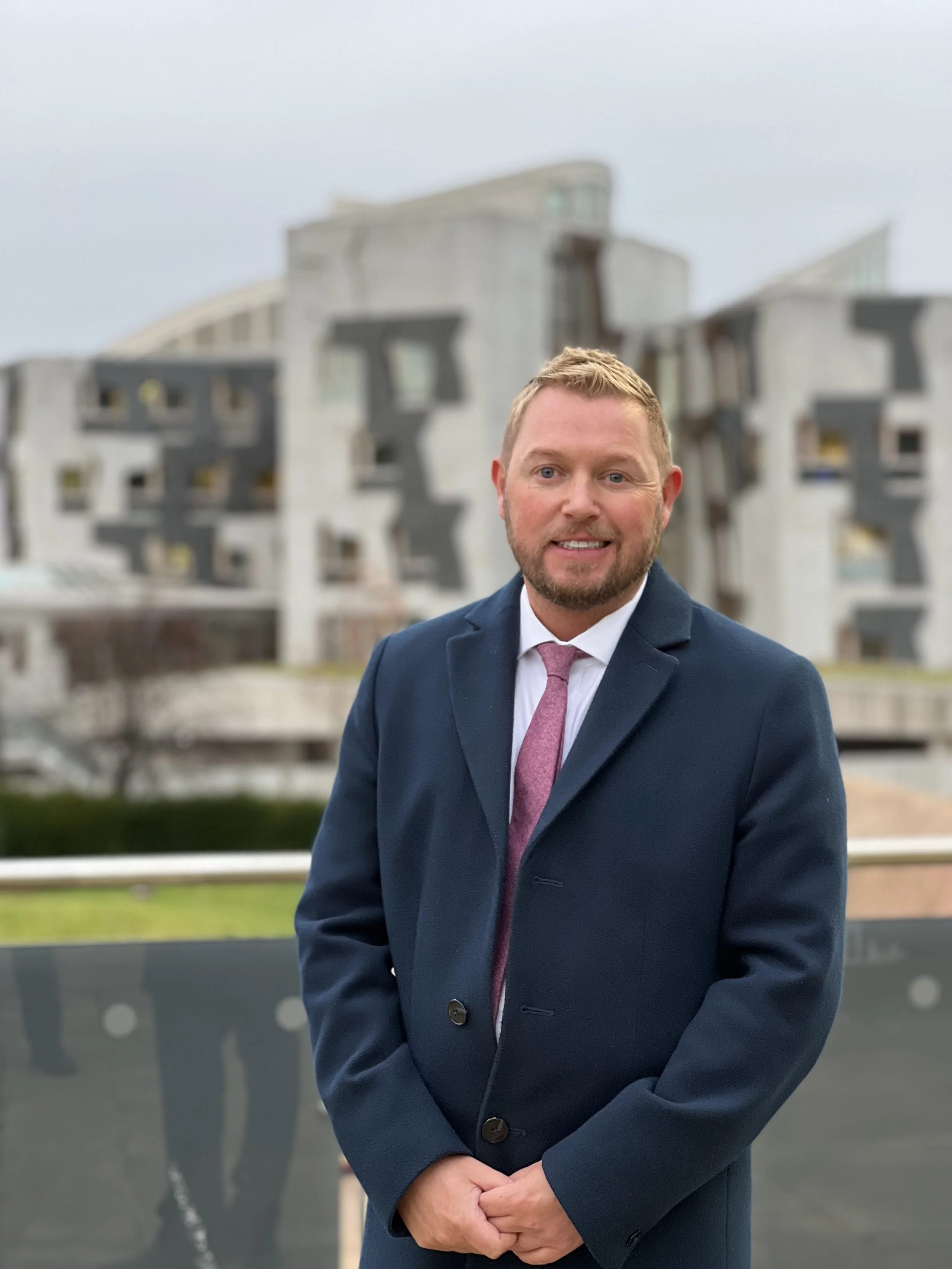 A man in a dark suit with a pink tie standing outdoors in front of a modern building with curved architecture. He is smiling slightly and has short blond hair and a beard.
