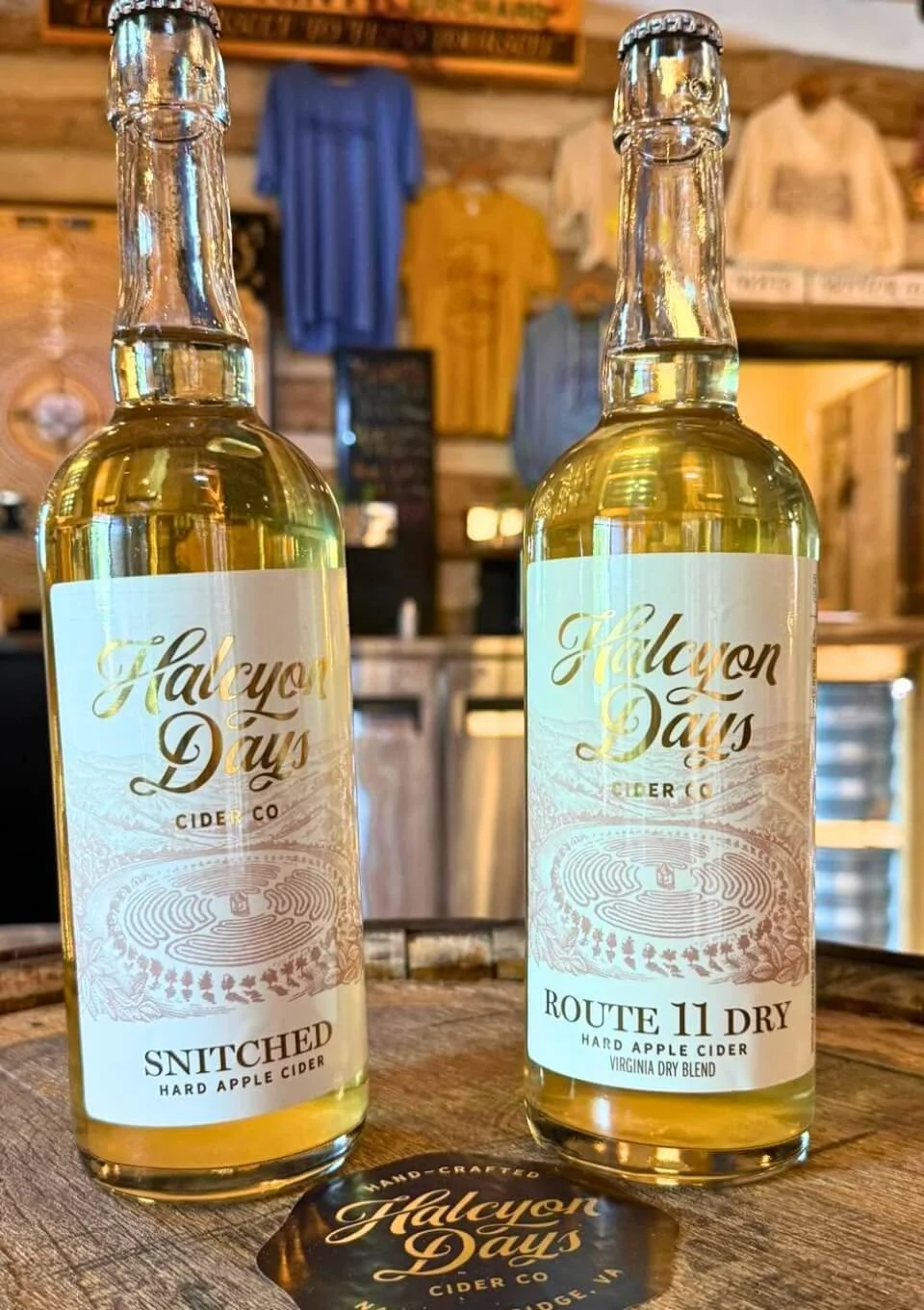 Two bottles of Halcyon Days hard cider on a wooden surface in the historic tasting barn. The left bottle is labeled 'Snitched Hard Apple Cider' and the right is 'Route 11 Dry Hard Apple Cider'.