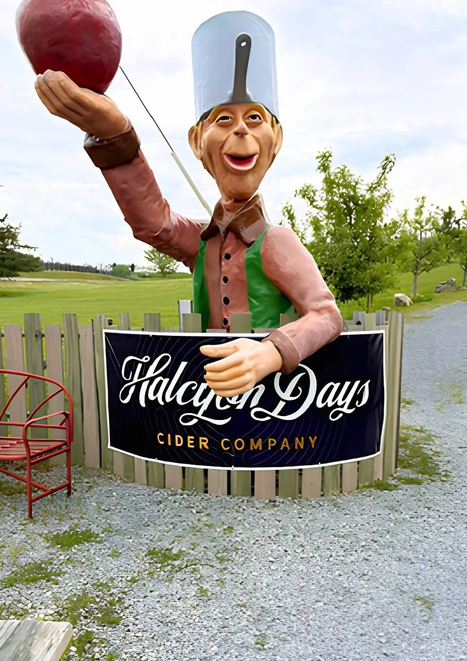 Large outdoor sculpture of a cheerful chef holding a red apple, with a sign for Halcyon Days Cider Company, set against a grassy area with trees and gravel pathway.