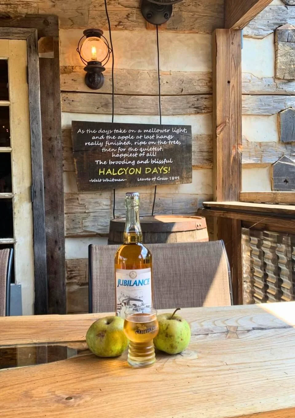 A wooden table with three apples, a small glass of hard apple cider, and a large bottle of hard cider. In the background, a wall with rustic wood planks and a decorative chalkboard sign with a quote about apple harvest and a glowing light fixture.