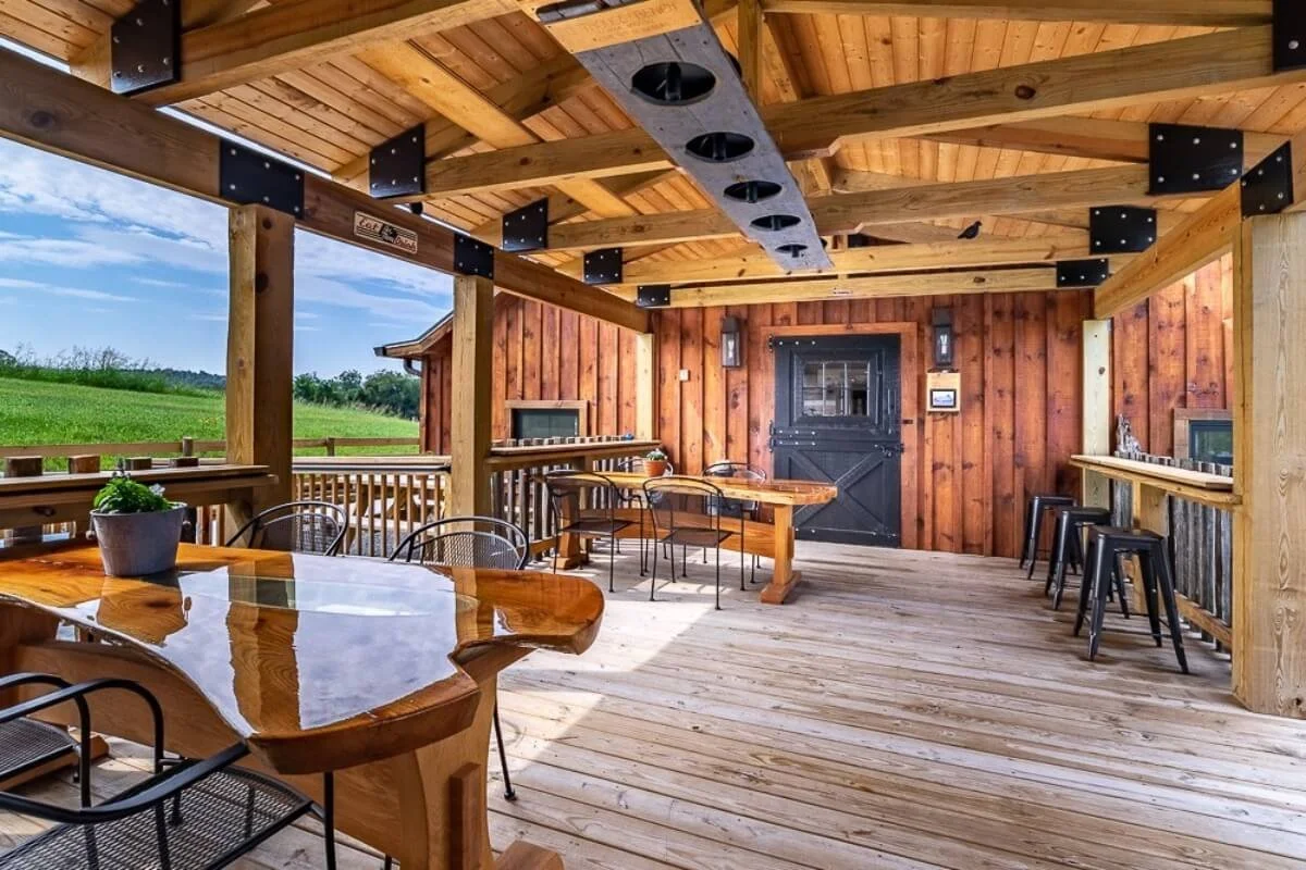 A covered outdoor patio with wooden flooring and walls, furnished with a large polished wood table, metal chairs, bar stools, and a black sliding barn door, overlooking a green field under a partly cloudy sky.