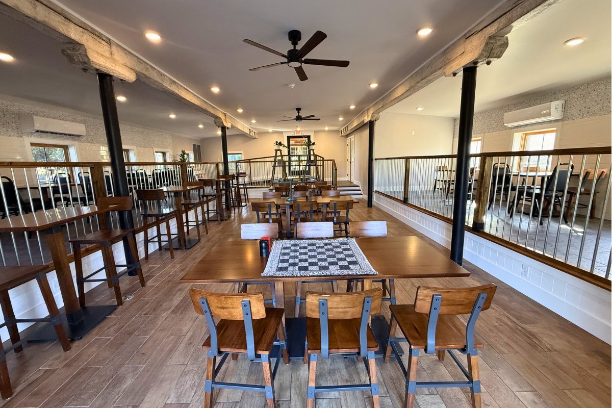 A covered outdoor patio with wooden flooring and walls, furnished with a large polished wooden table, black metal chairs, and barstools. The patio has a roof made of wooden beams and is open on the sides, overlooking a grassy landscape under a blue sky.