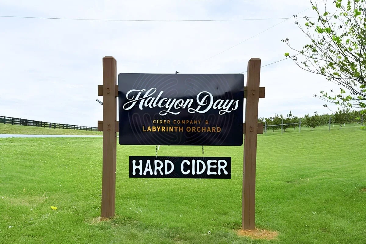 Sign for Halcyon Days Cider Company & Labyrinth Orchard advertising hard cider, set on a grassy field with a tree on the right.