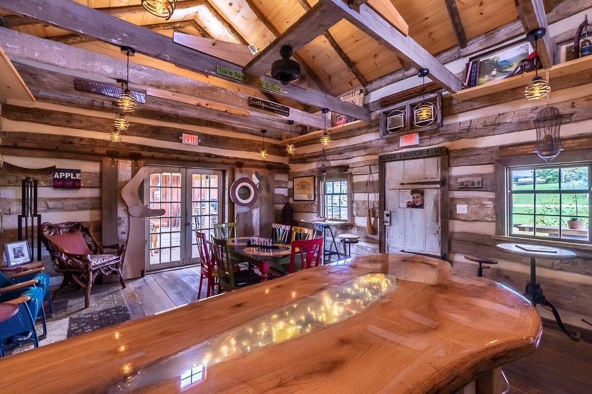 Interior of historical tasting barn with a curved wooden bar in the foreground, featuring string lights embedded in its surface. The walls are made of reclaimed wood planks, decorated with art, signs, and photos.