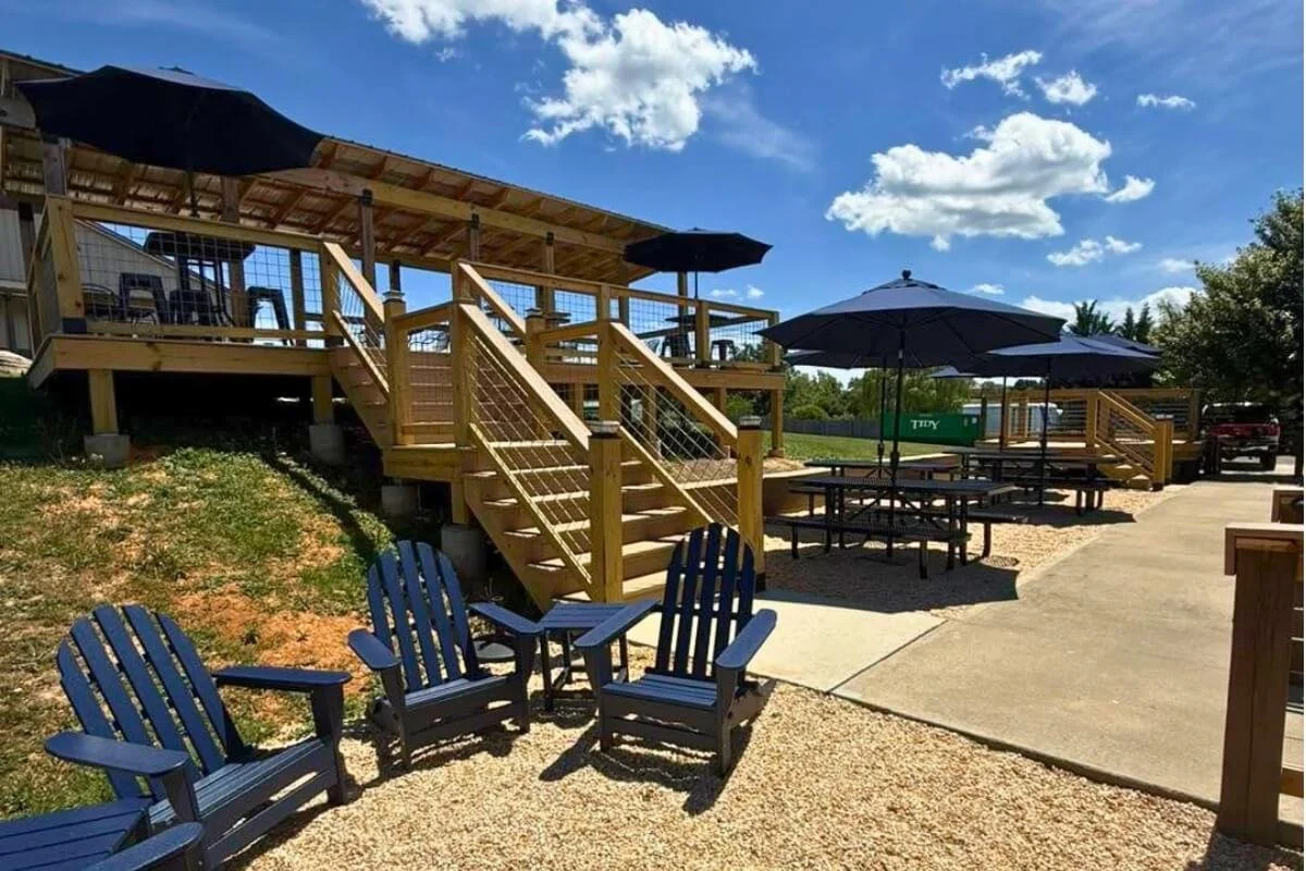 Outdoor seating area with wooden staircase, picnic tables with umbrellas, Adirondack chairs, and a gravel pathway under a partly cloudy sky.