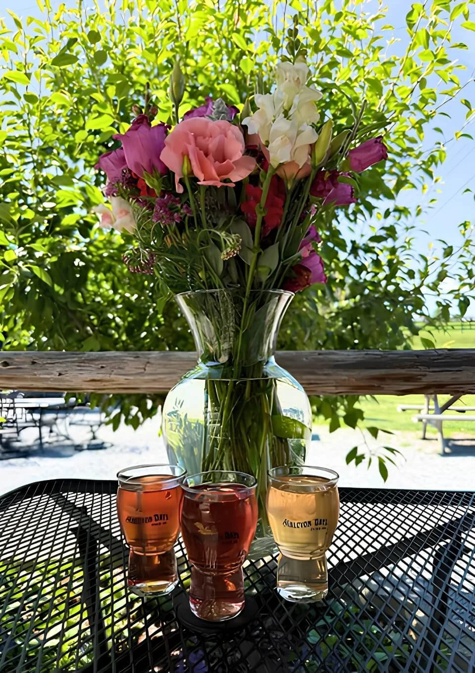A glass vase with a bouquet of pink, white, and purple flowers on an outdoor table, with three glasses of hard cider in front, and green foliage in the background.