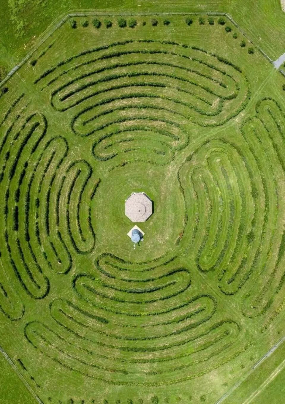 Aerial view of the Halcyon Days Cider Company's Labyrinth Orchard with concentric pathways and a small octagonal structure at the center, surrounded by grass and trees.