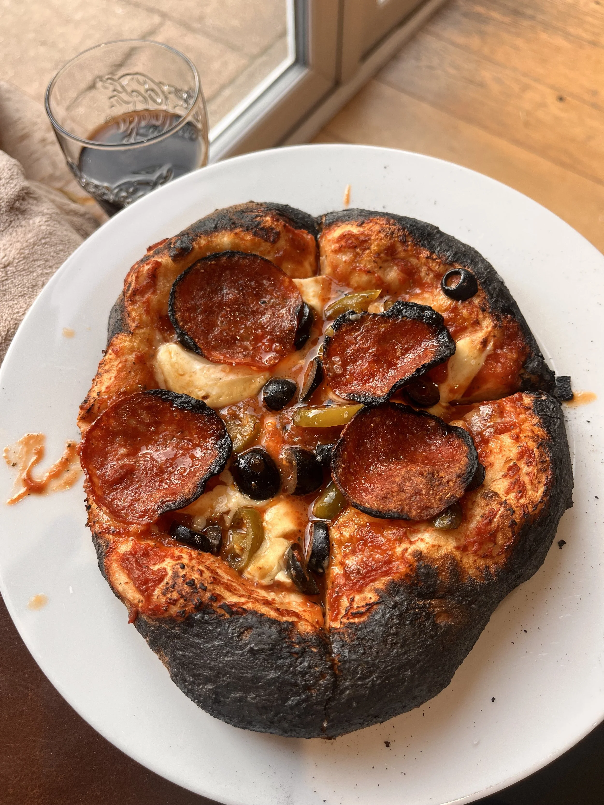 Blackened pizza topped with pepperoni, black olives, and green peppers on a white plate, with a glass of soda and a window in the background.
