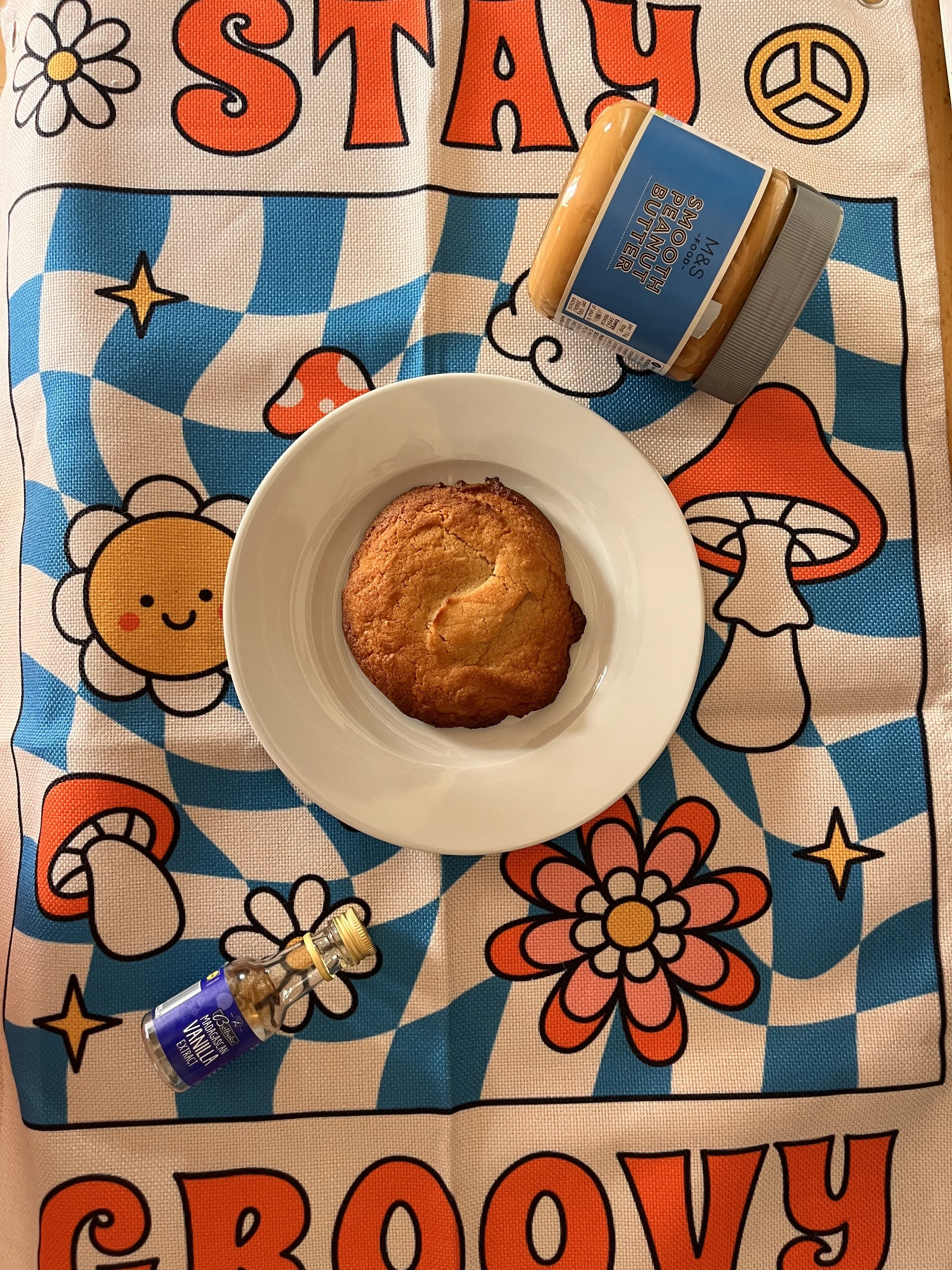 A white plate with a freshly baked muffin sits on a colorful fabric with cartoon flowers, mushrooms, and peace symbols. A jar of banana bonito and a small bottle are also in the scene.