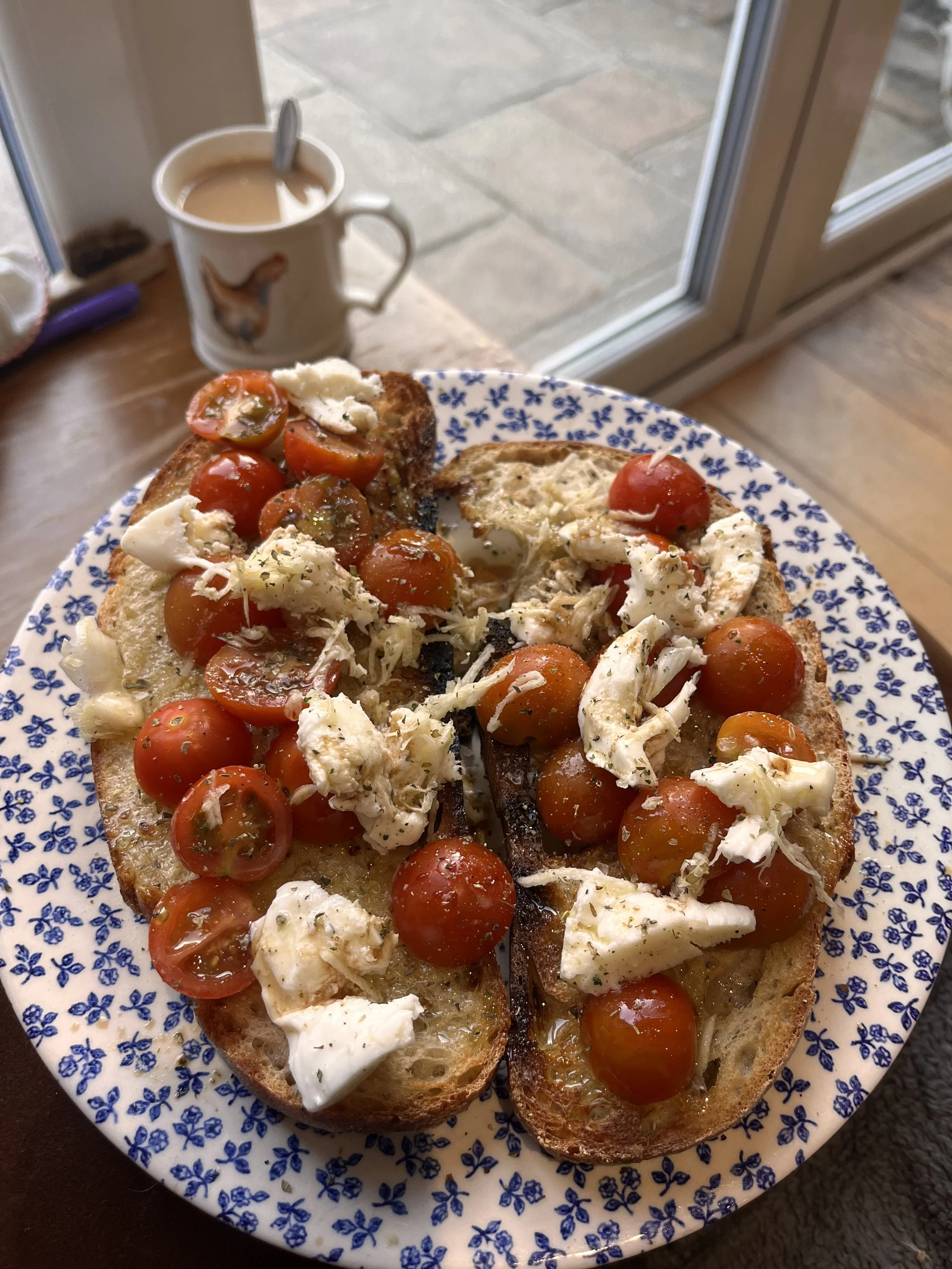 Open-faced bread topped with cherry tomatoes, shredded cheese, and cream cheese, served on a blue floral plate. A mug of coffee with a spoon is in the background near a window.
