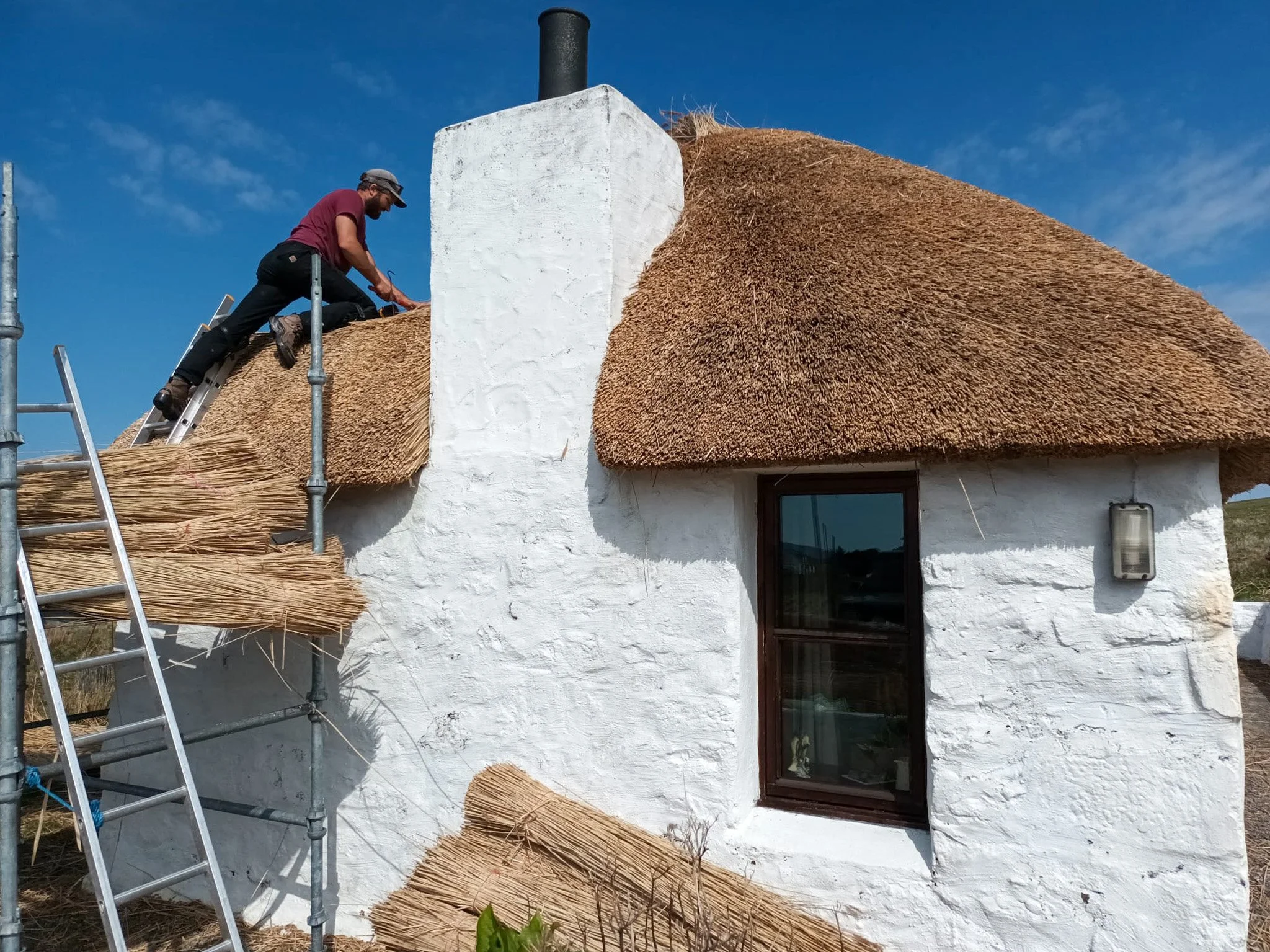 A person working on thatched roofing of a white cottage with a chimney, using scaffolding and a ladder under a blue sky.