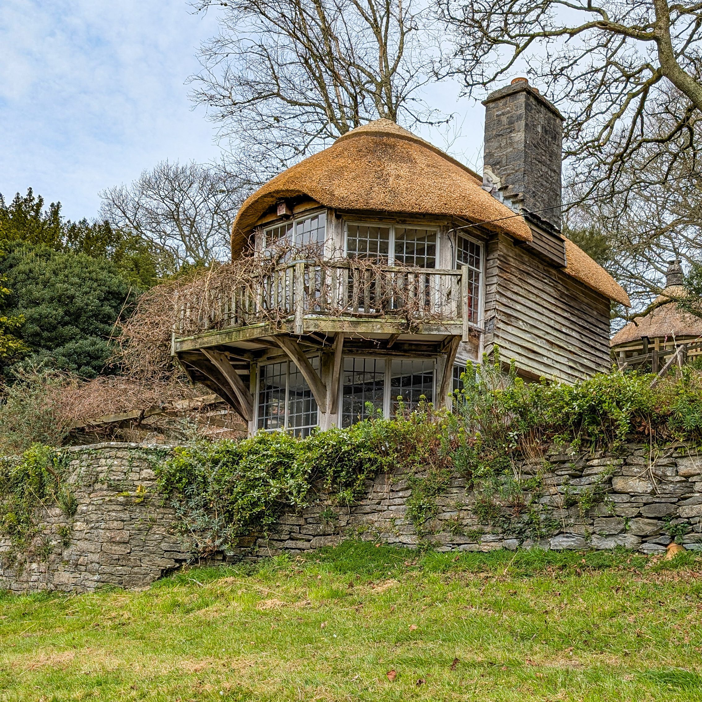 A house with a thatched roof, large windows, and a chimney, surrounded by trees and a brick wall with greenery in the foreground.