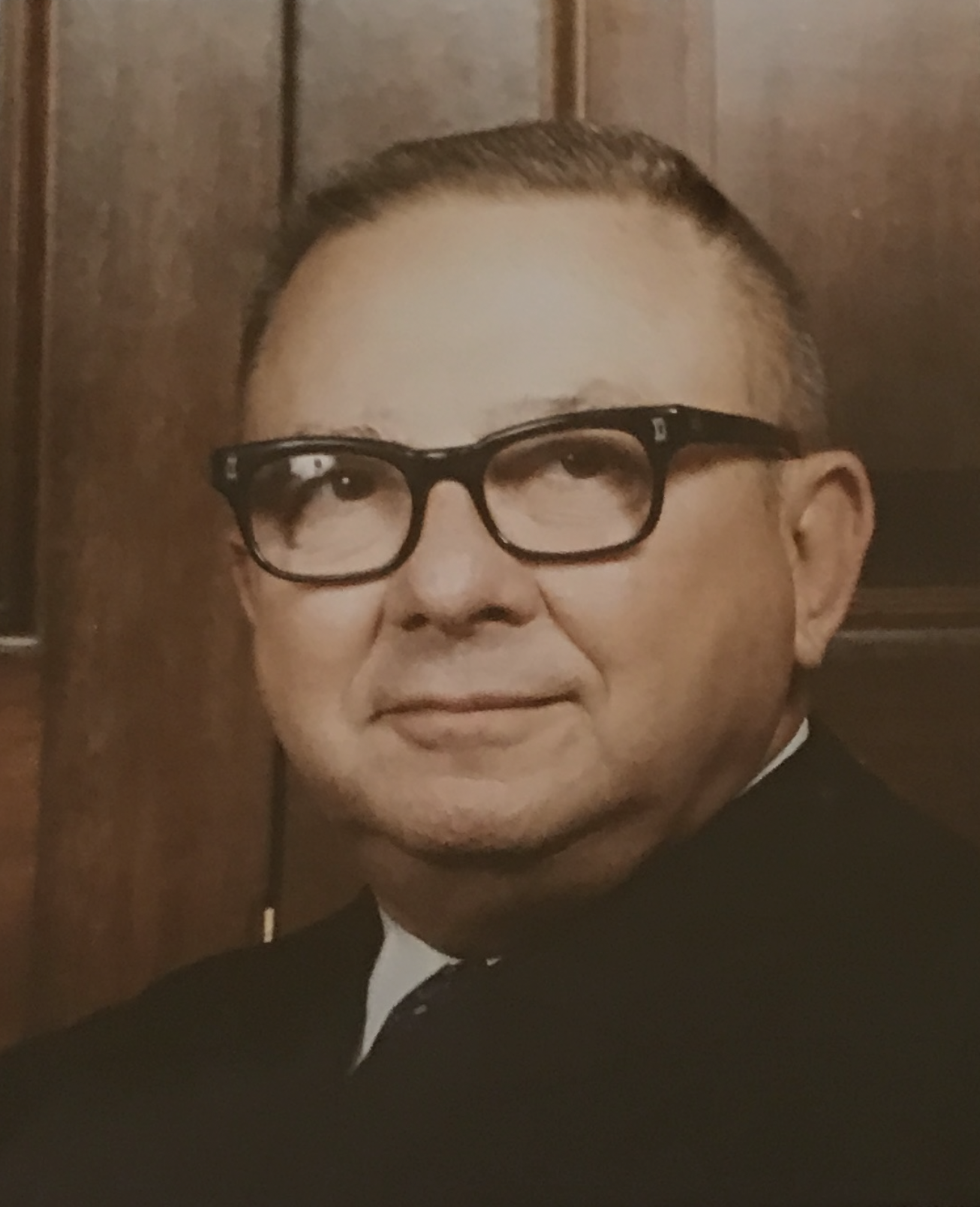 Headshot of a man with short hair, glasses, and a slight smile, dressed in formal attire, standing in front of a wooden background.