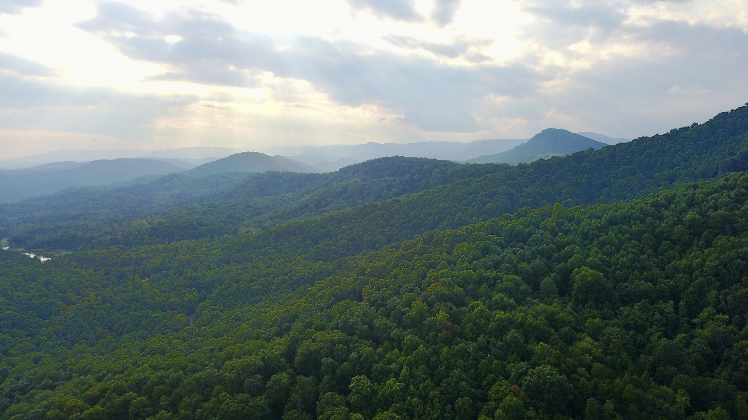 A landscape view of a lush, green mountain range with rolling hills under a partly cloudy sky with sunlight filtering through the clouds.