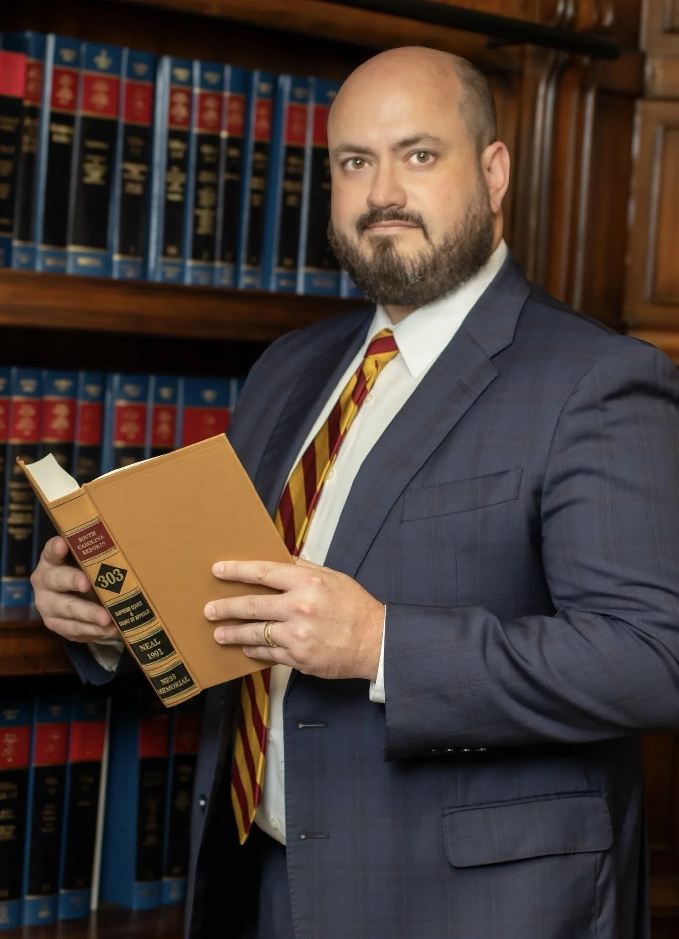 A man in a dark suit and striped tie holding an orange law book in a library with legal bookshelves in the background.