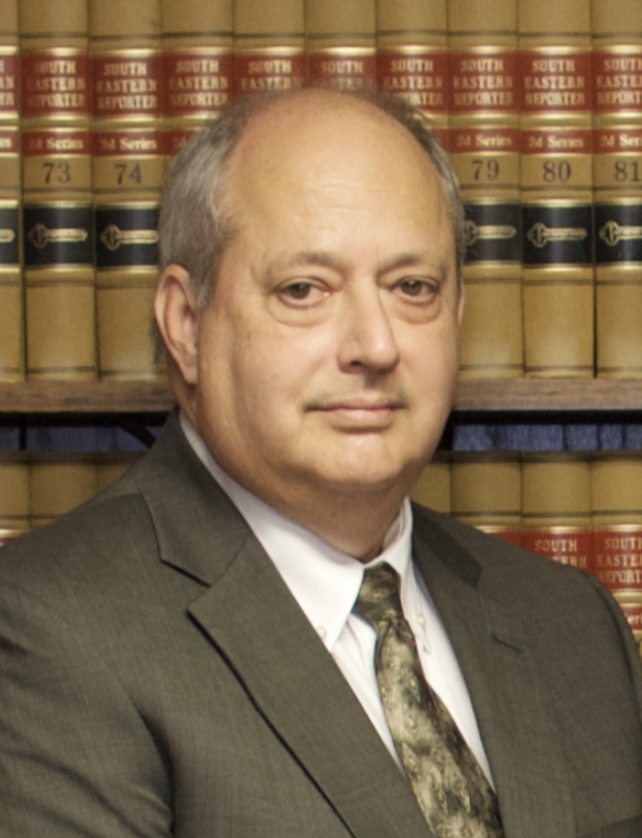 A middle-aged man in a suit with a patterned tie, standing in front of a bookshelf filled with law books.